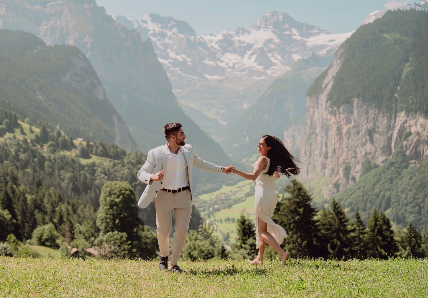 Joyful couple holding hands and running through a sunlit Alpine meadow with majestic snow-capped mountains in background.
