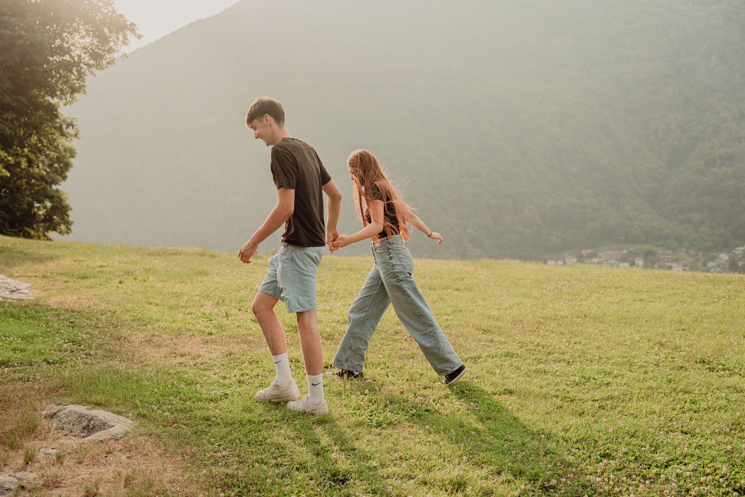 Couple walking hand-in-hand across a vast green alpine meadow with misty mountains behind them.