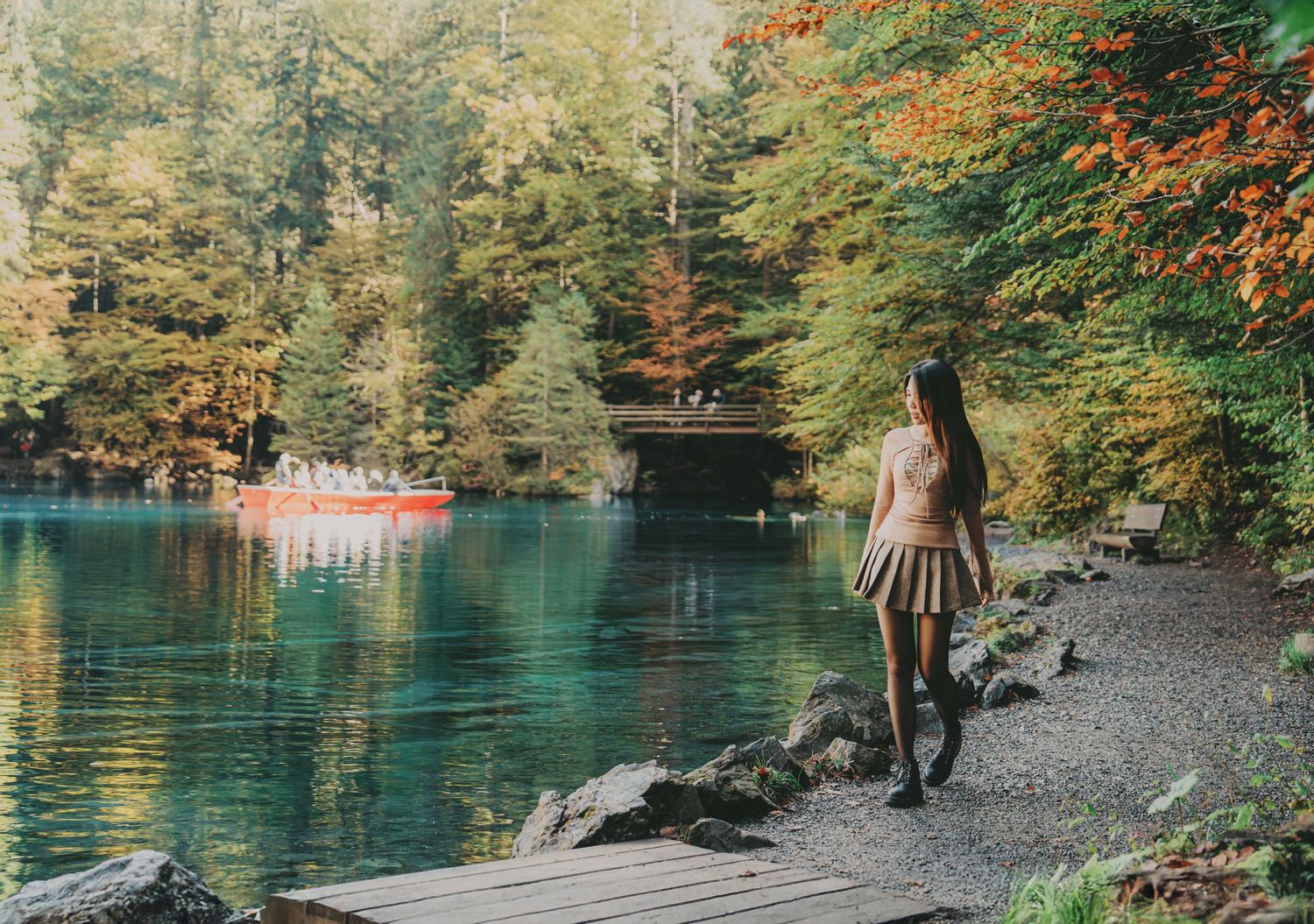 Woman in skirt stands on wooden dock overlooking a turquoise lake with red boat and autumn foliage.