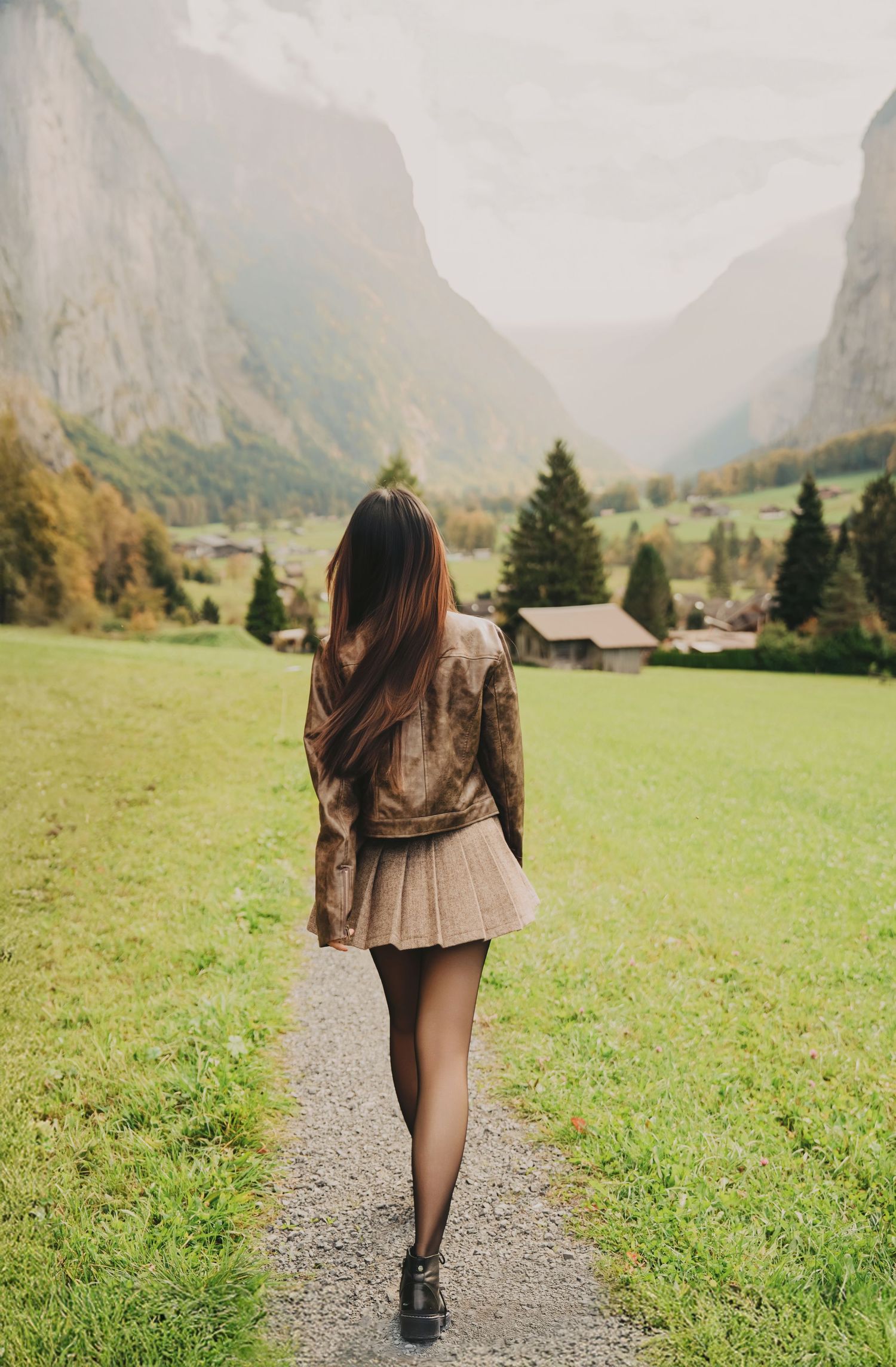 Woman with long hair in brown jacket and skirt walking away on path in Swiss alpine valley.