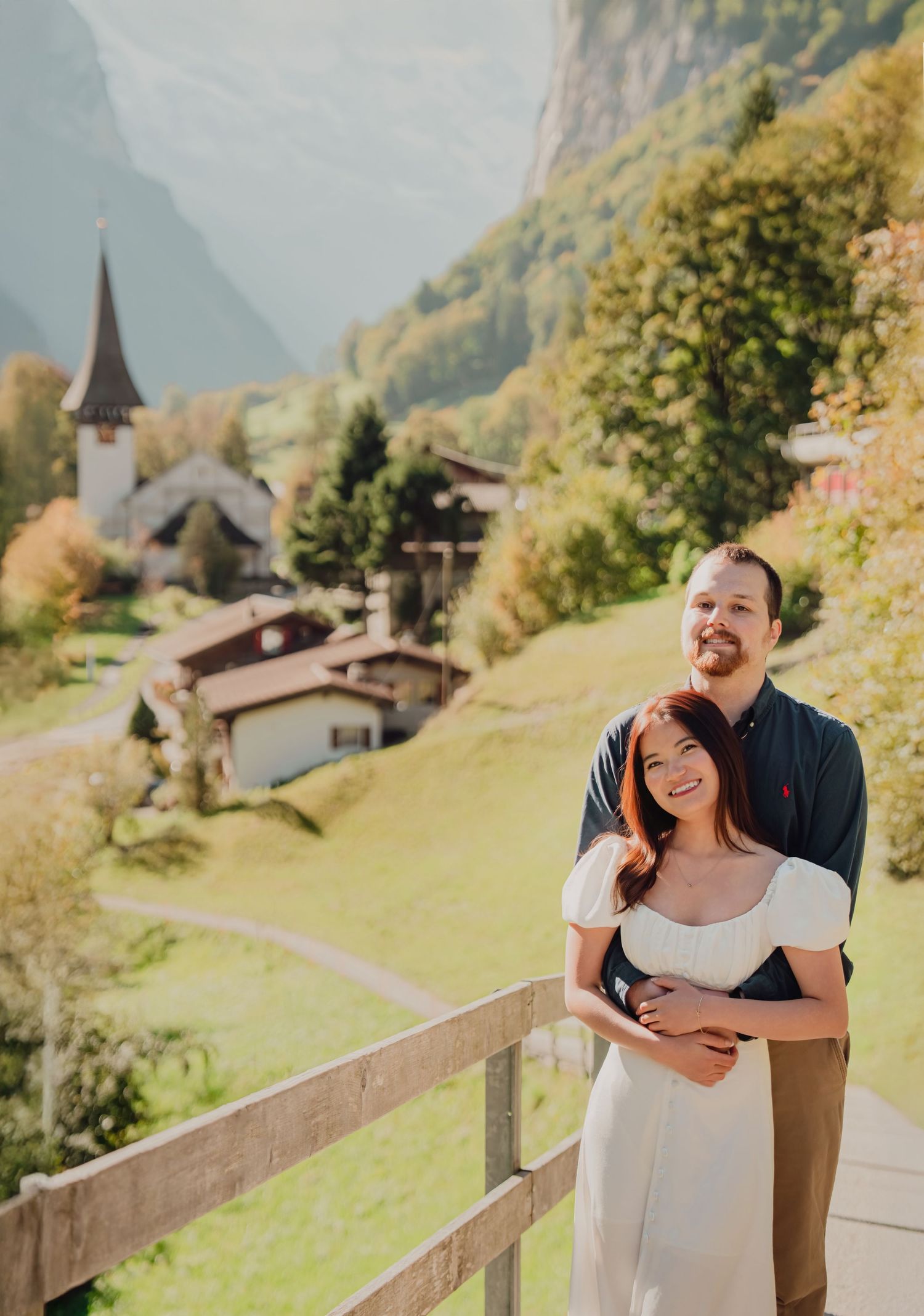 A smiling couple embracing on a balcony with a stunning Swiss alpine village and mountains behind them.