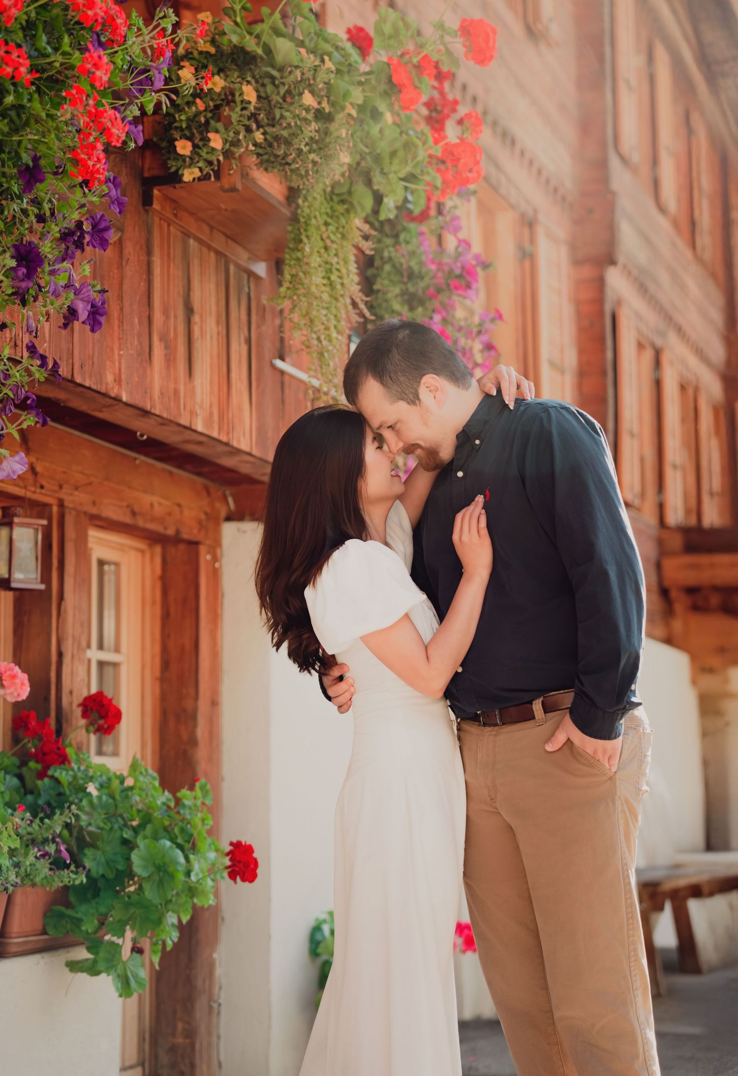 Couple sharing a romantic kiss beside colorful hanging flower baskets on a charming rustic wooden building exterior.