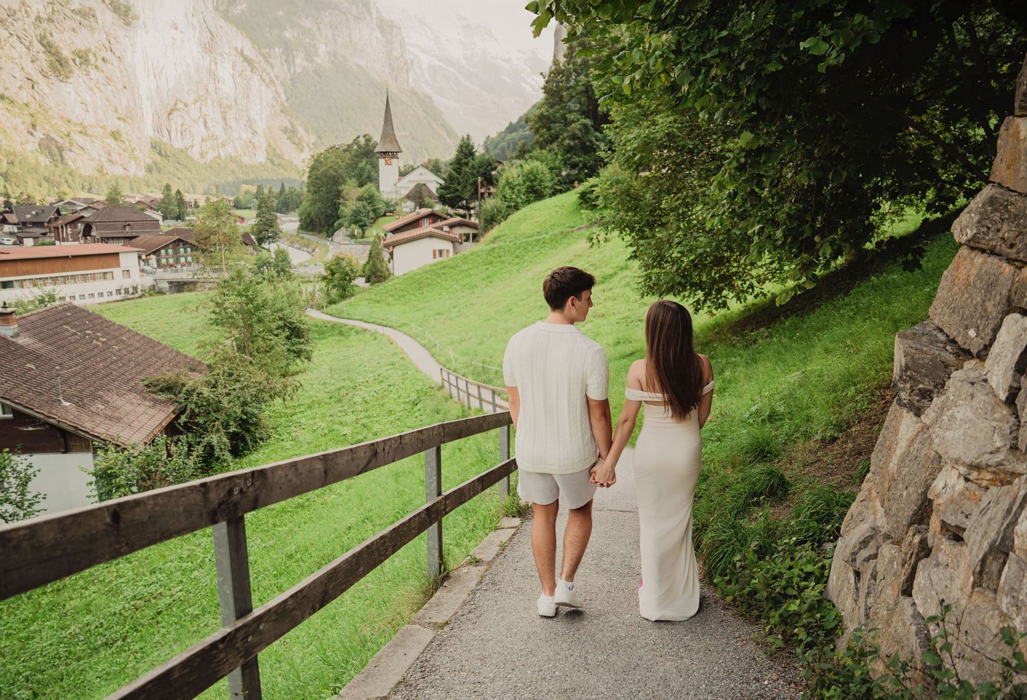 Couple holding hands walking down a path in a scenic Swiss alpine village with a church visible.