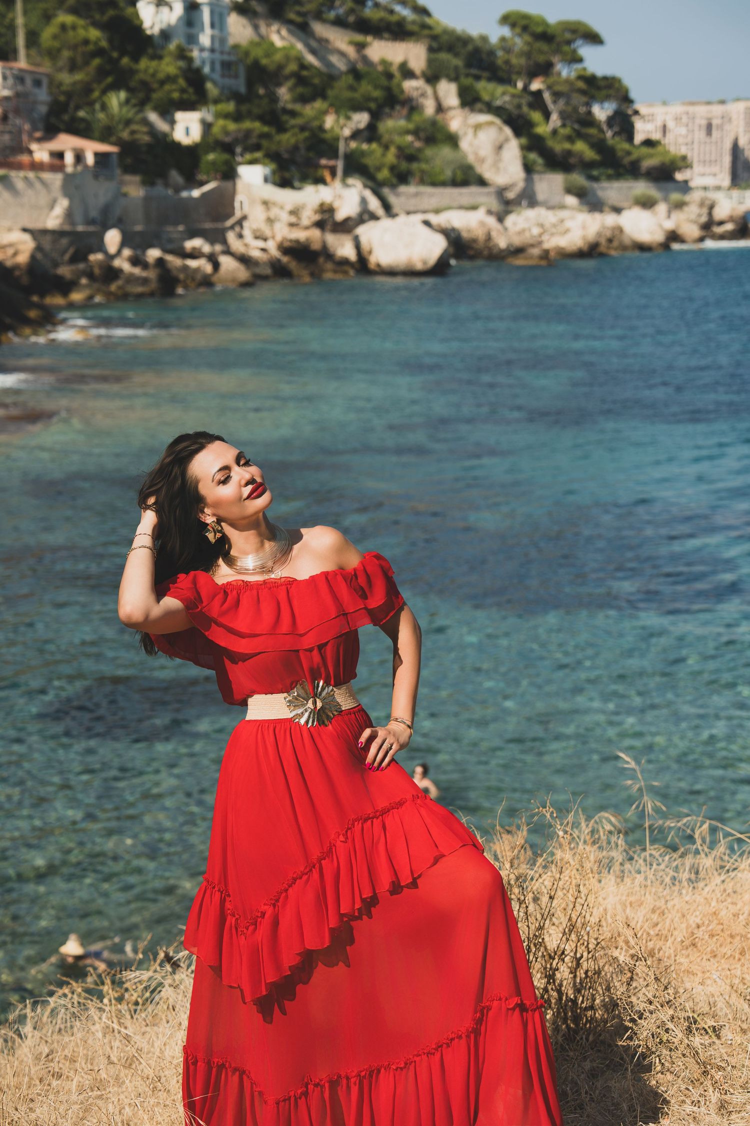 Woman in red ruffled two-piece dress laughing on rocky coast with turquoise Mediterranean waters.