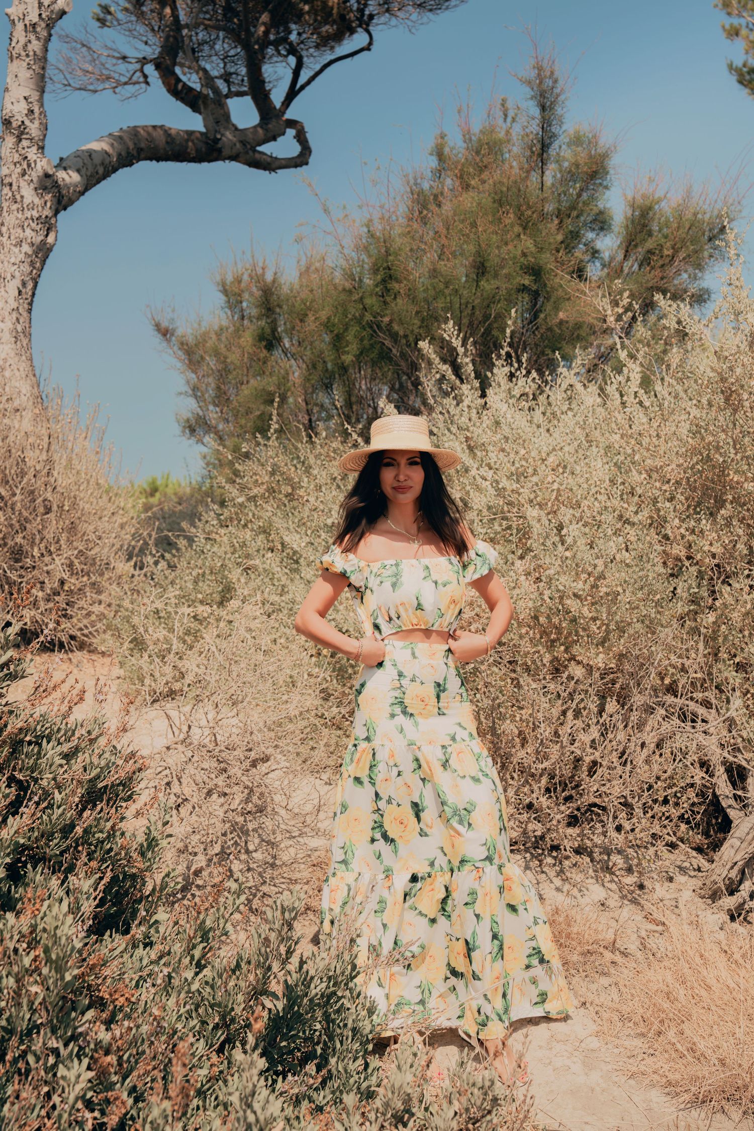 Woman in yellow floral off-shoulder dress and straw hat standing in dry coastal shrubbery.
