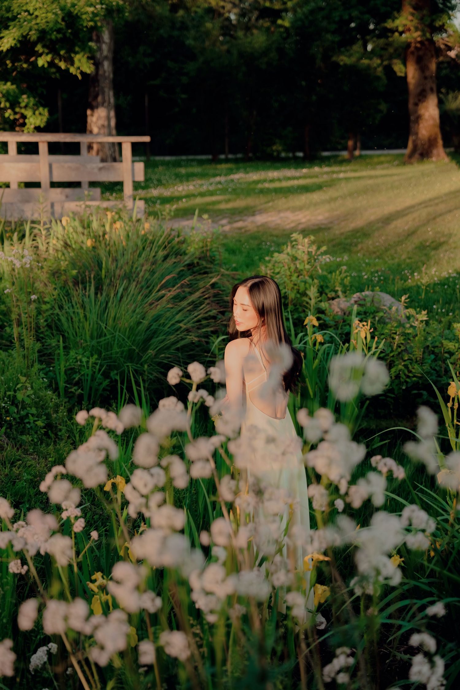 Young woman in white dress standing among wildflowers in a sunlit garden at golden hour.