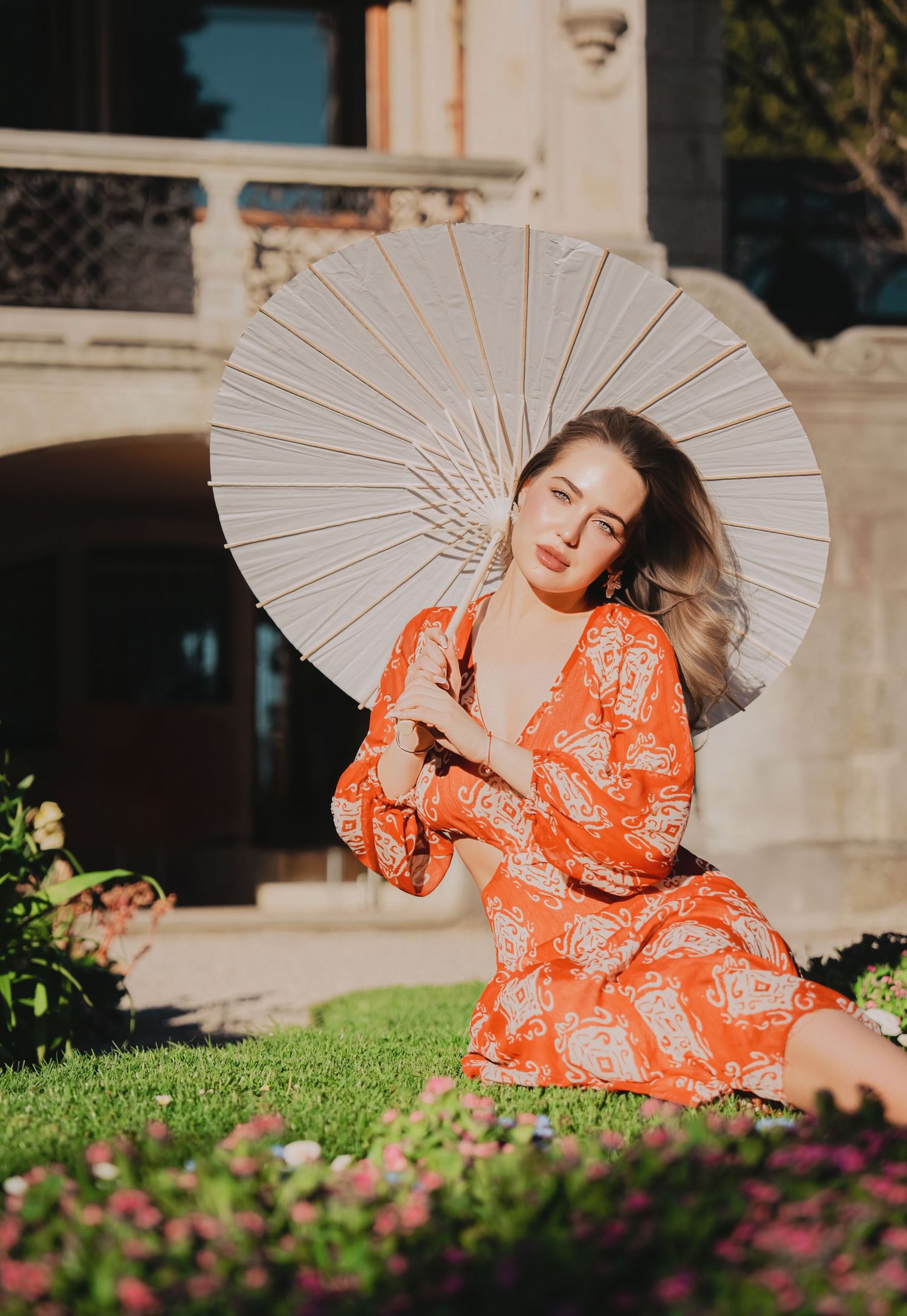 Woman in orange patterned dress sitting on grass holding white parasol umbrella outdoors.