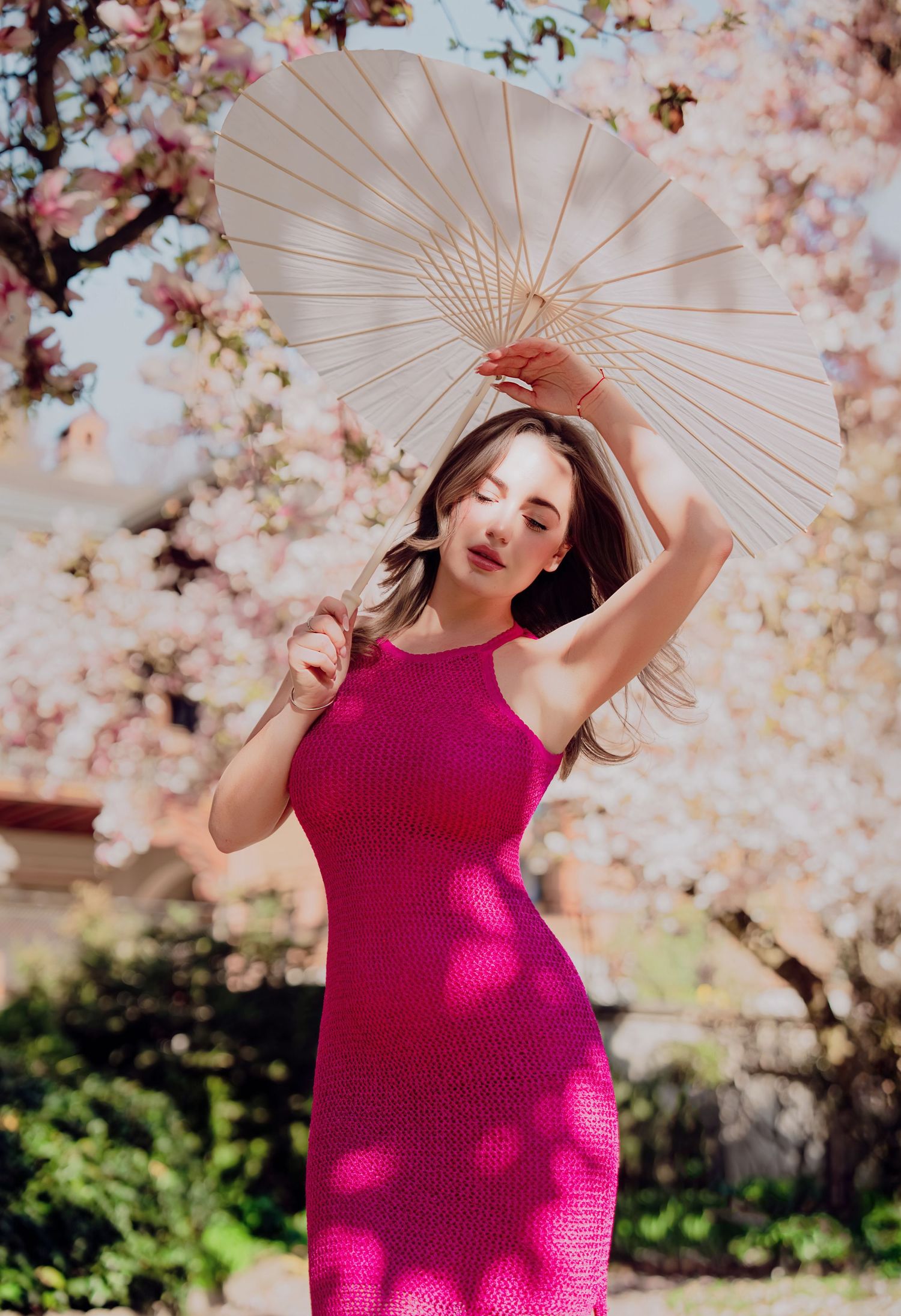 Woman in hot pink knit dress holding white parasol under blooming cherry blossom trees.