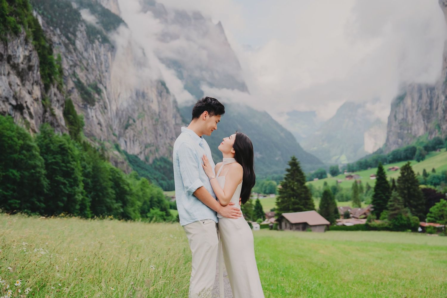Couple embracing in a lush green meadow with dramatic misty mountains and pine trees in the background.