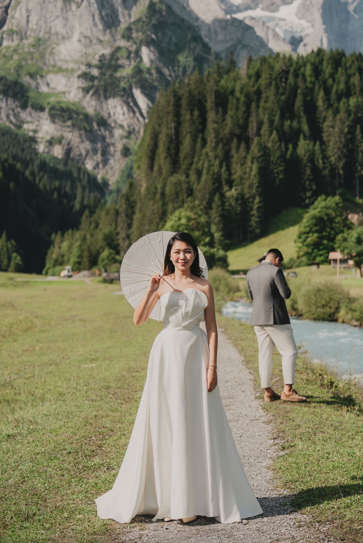 Smiling bride in elegant white dress holds white parasol on scenic mountain valley path in Switzerland.