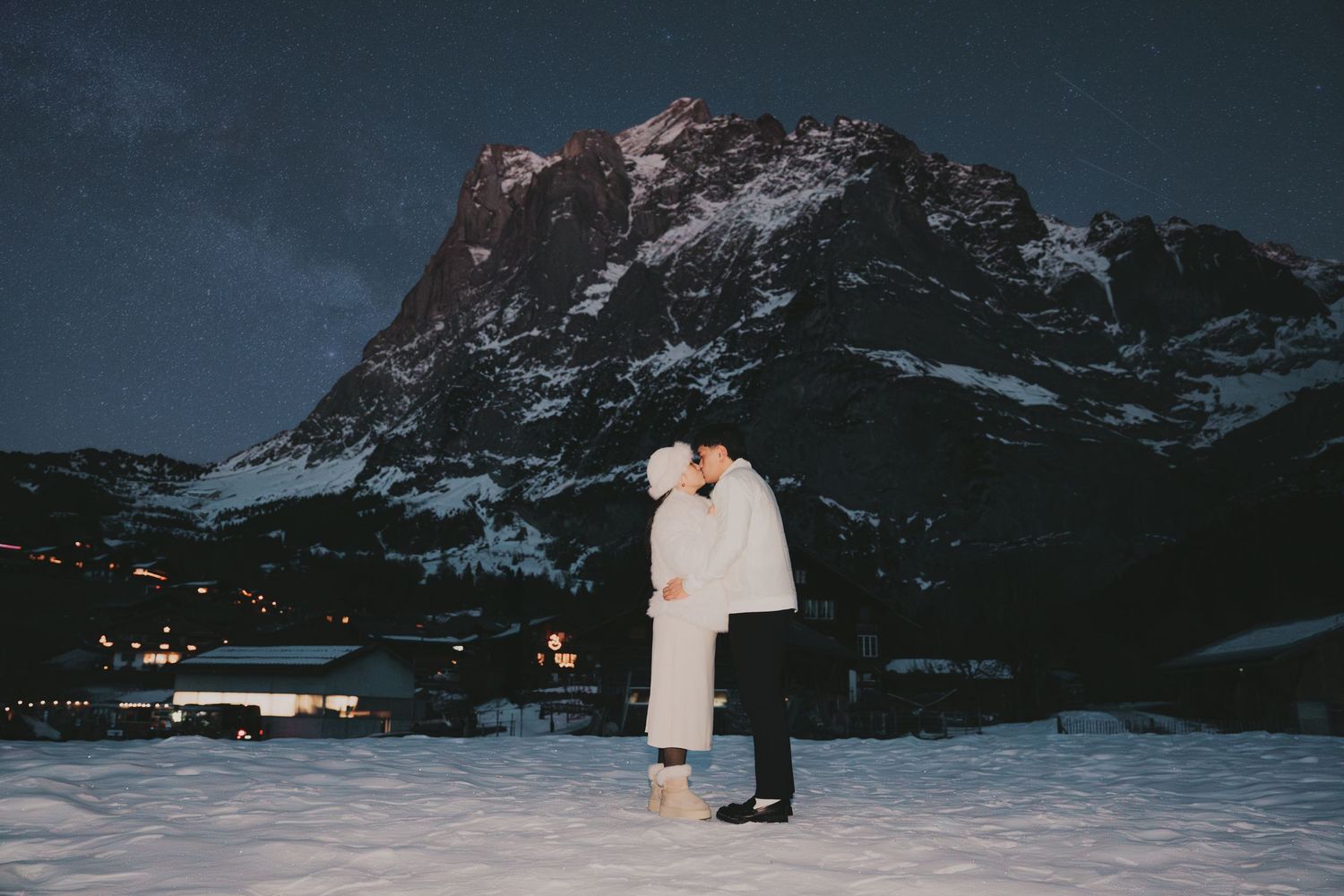 Romantic couple kissing under starry night sky in snowy alpine village with illuminated mountain backdrop.