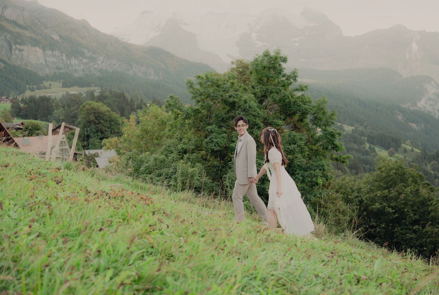 Bride and groom walking hand in hand up lush green Swiss alpine hillside with mountain scenery.