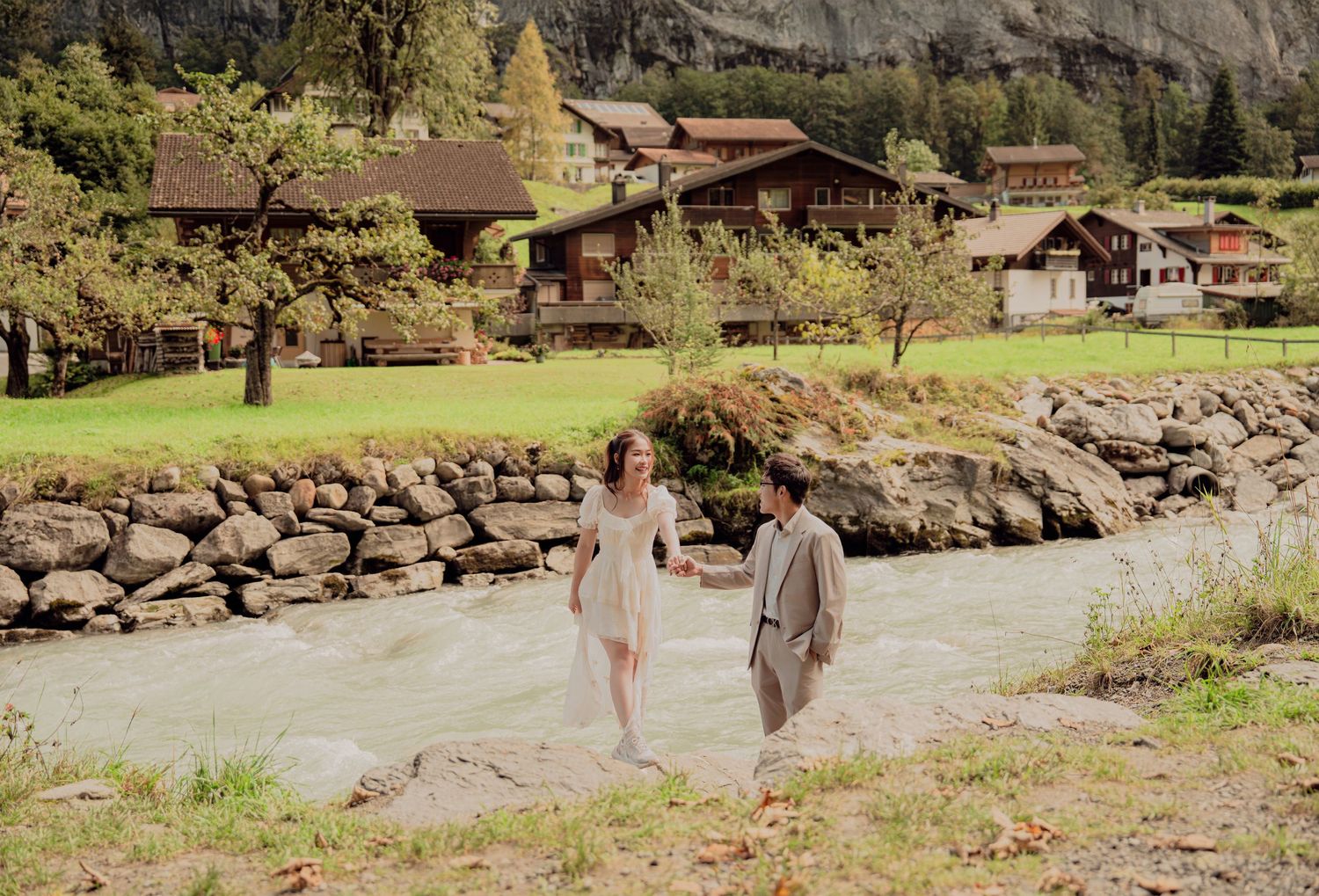 Couple walking hand in hand along a riverbank with lush green Swiss alpine village in the background.