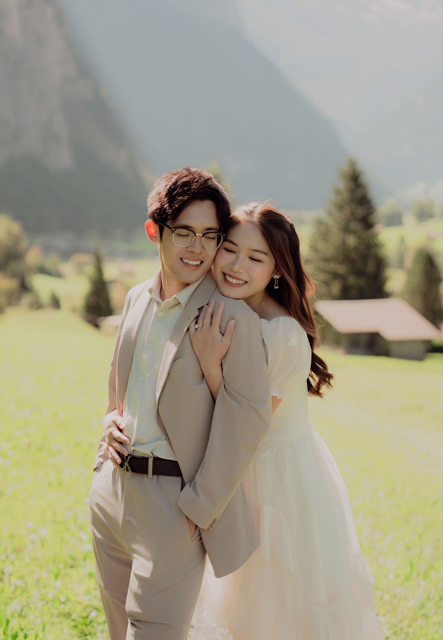 Couple embracing in a green meadow with Swiss Alps and pine trees in the background.