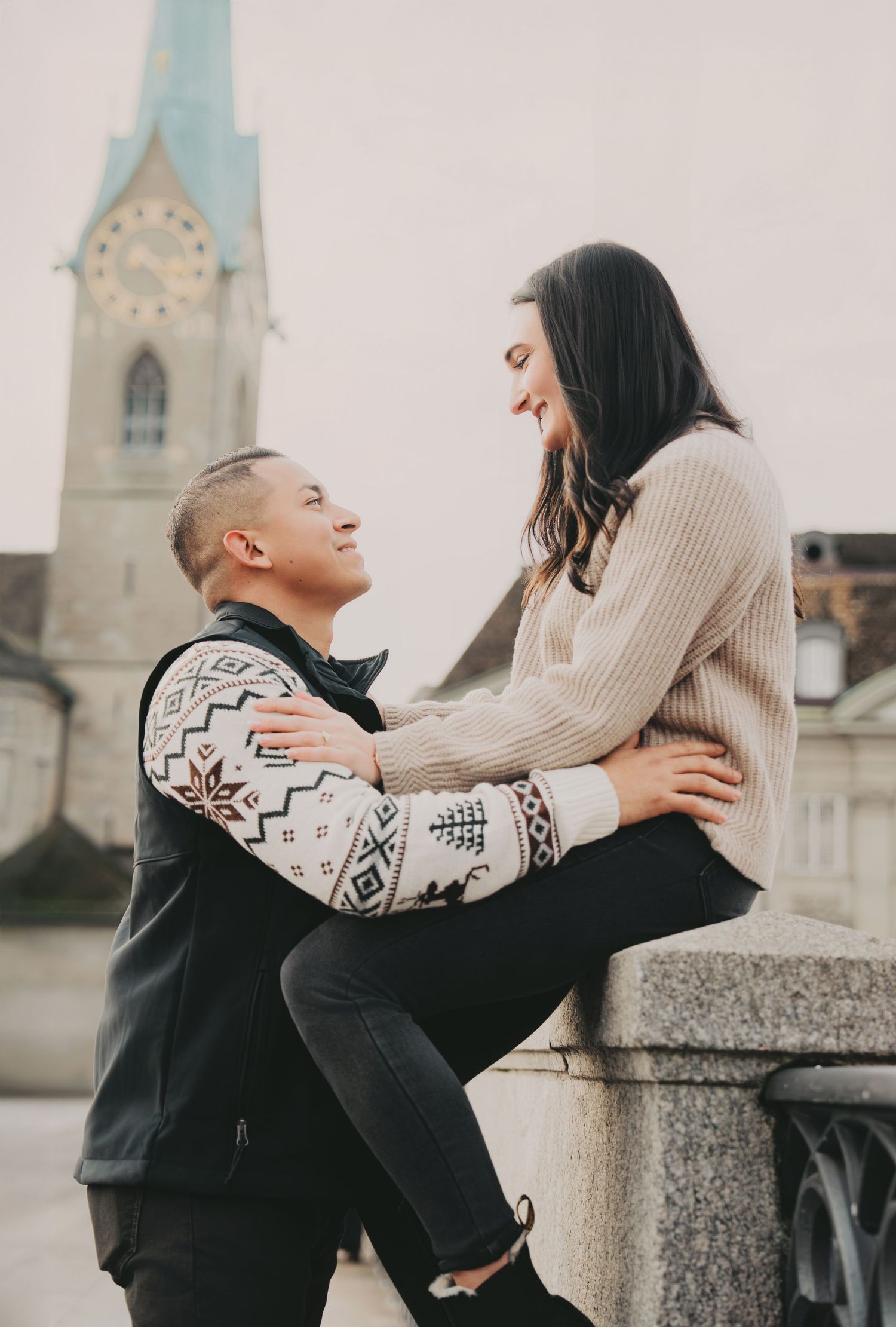 Couple laughing together on a stone ledge with Fraumunster Church tower in Zurich backdrop.