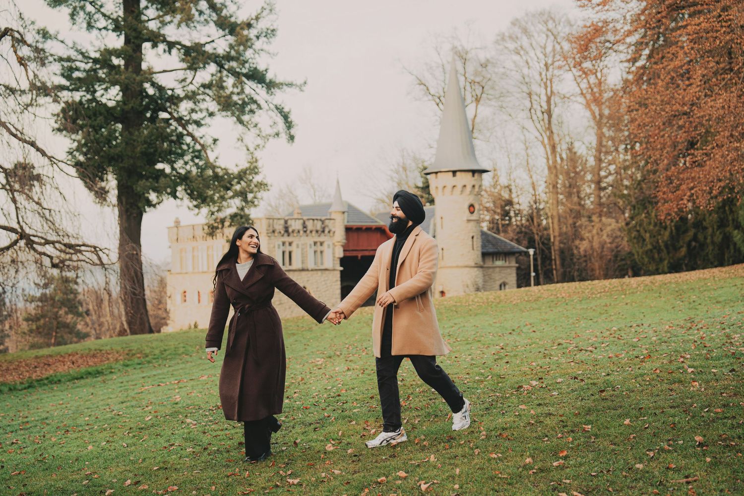 Engaged couple walking hand in hand on grass with a historic castle backdrop in fall season.