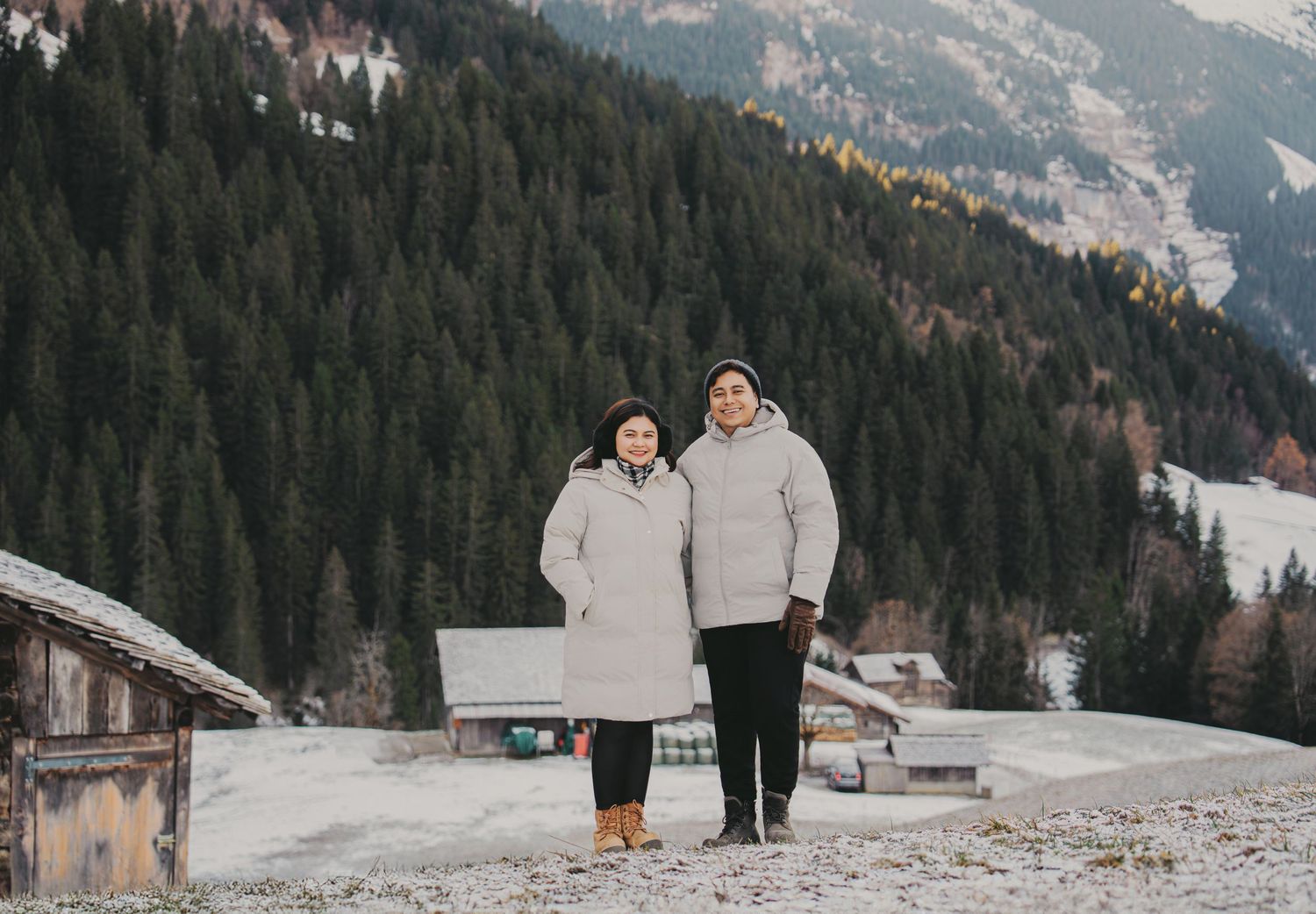 Smiling couple in matching white coats walking through frost-covered alpine meadow with mountain views.