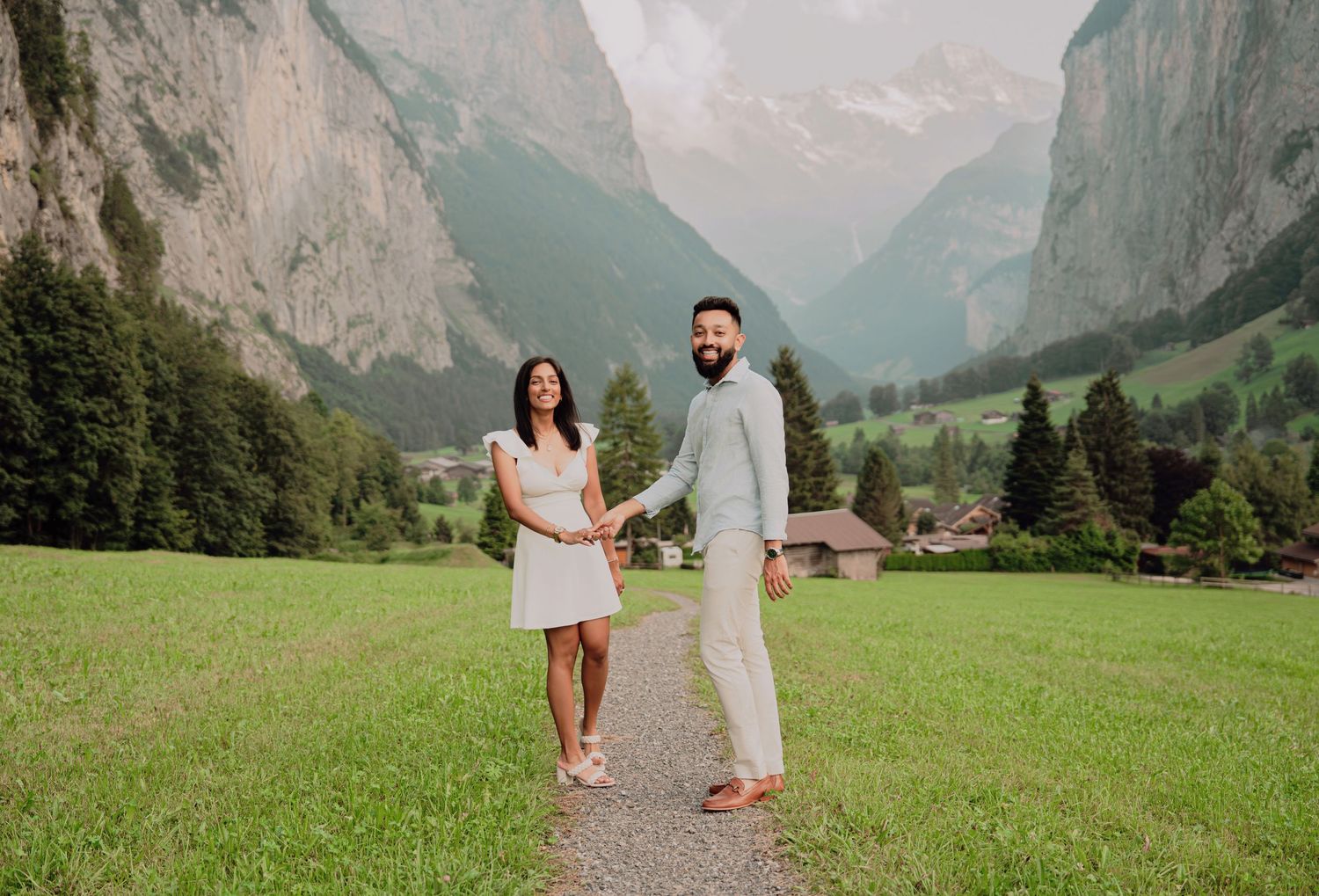 Couple holding hands on a scenic gravel path in a lush Swiss Alpine valley with tall mountains.