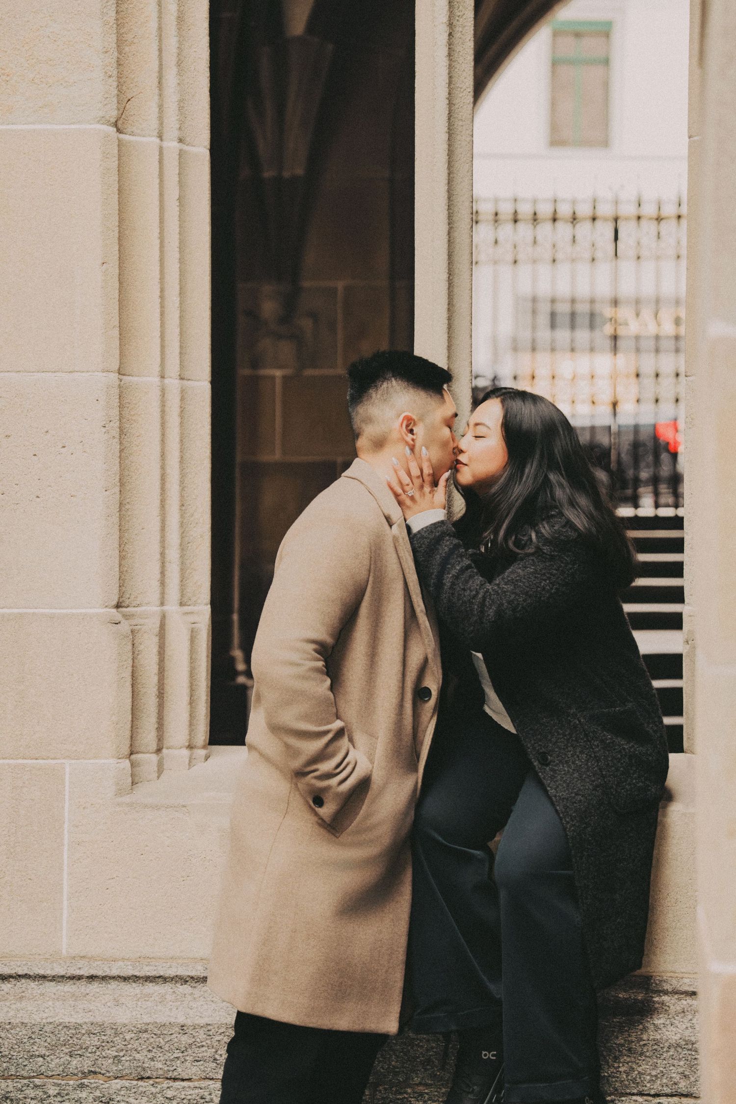Couple sharing a kiss beside stone columns outside a historic Gothic stone building.