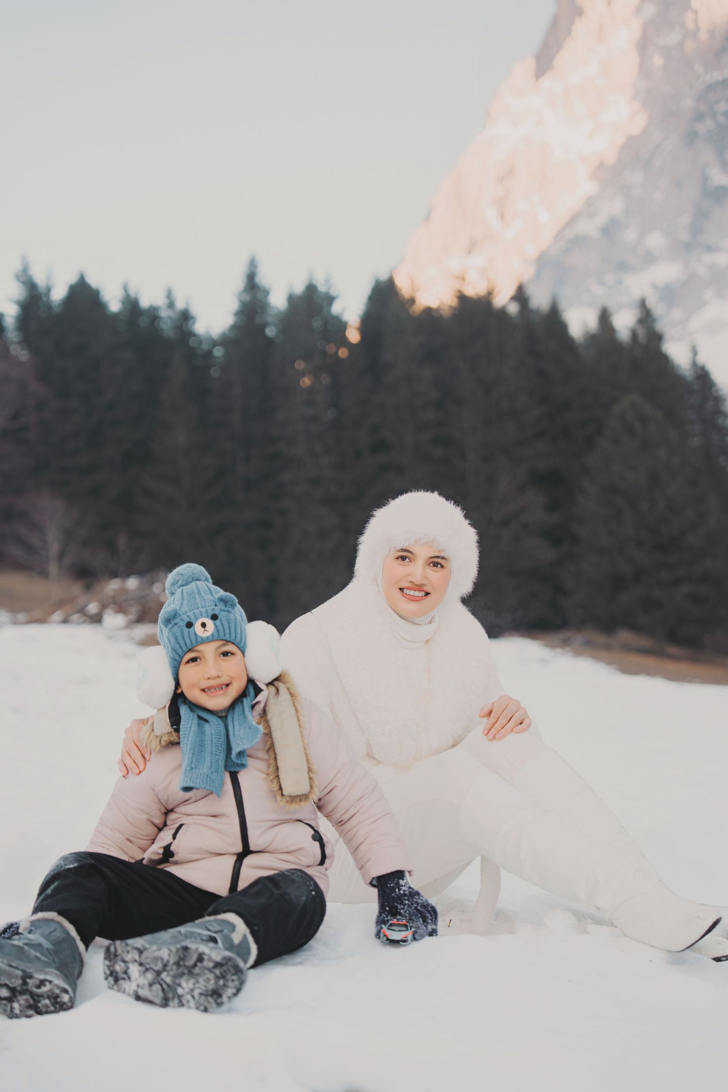Smiling mother and daughter posing together in snowy alpine setting with pine trees and mountain peak.