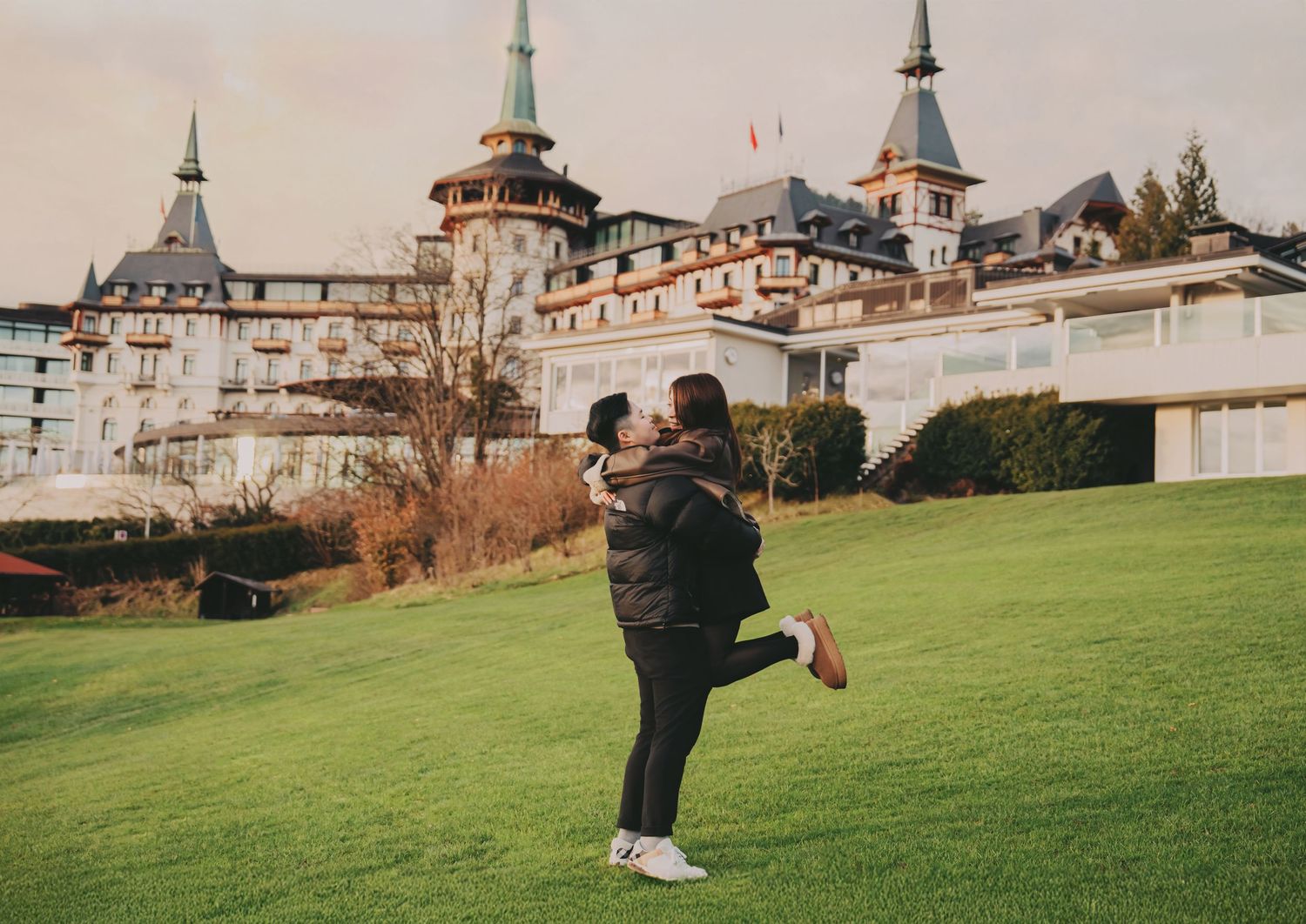 Man lifting woman joyfully in front of grand European castle on lush green grounds.