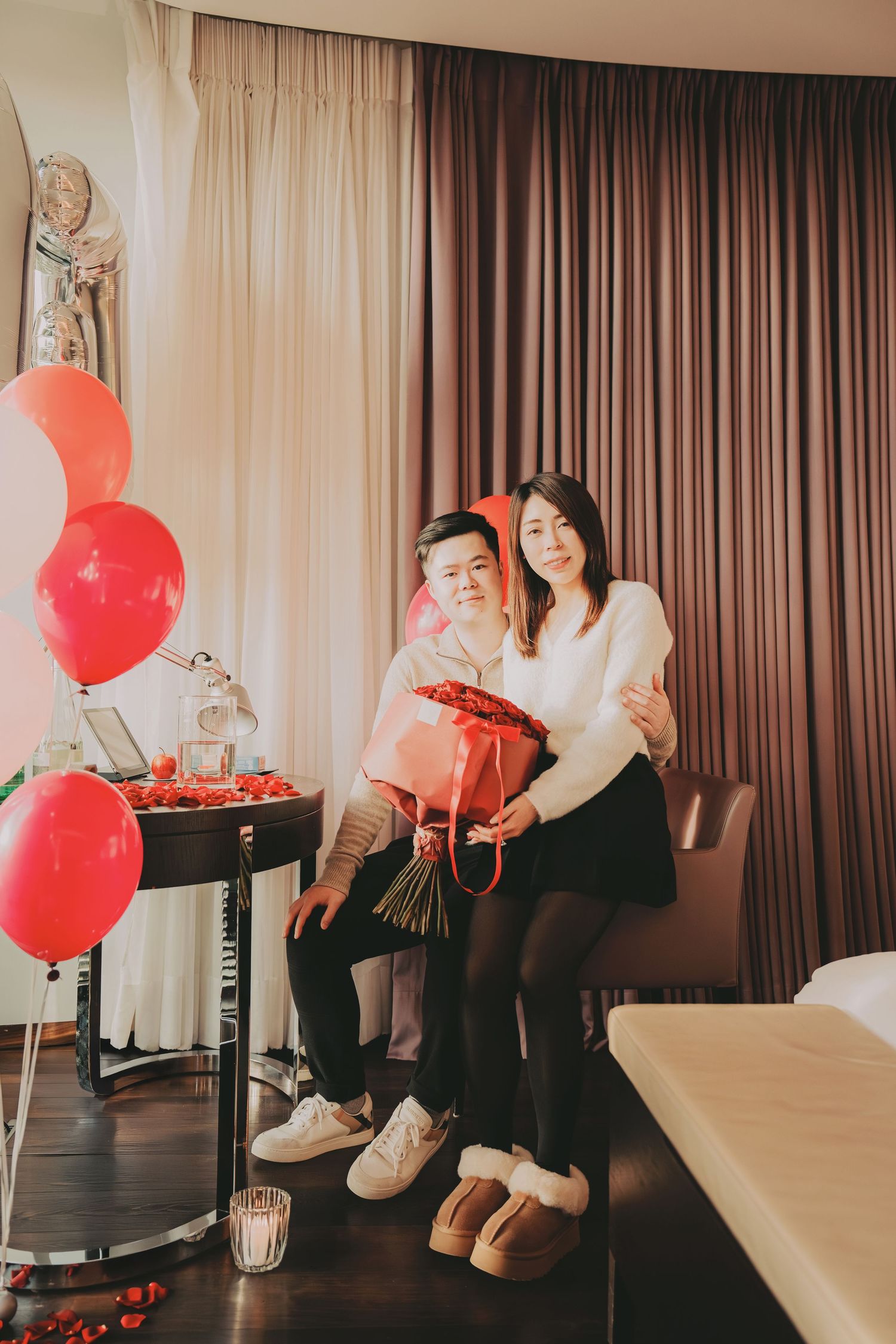 Asian couple posing with red roses and heart balloons in a romantic hotel room Valentine's Day setup.