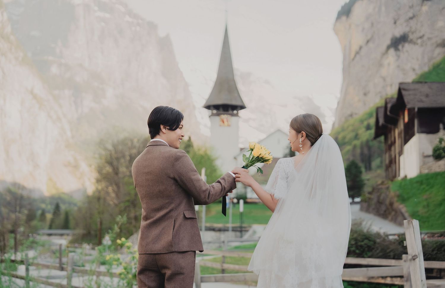 Groom presenting yellow bouquet to bride in white veil with a Swiss alpine church backdrop.