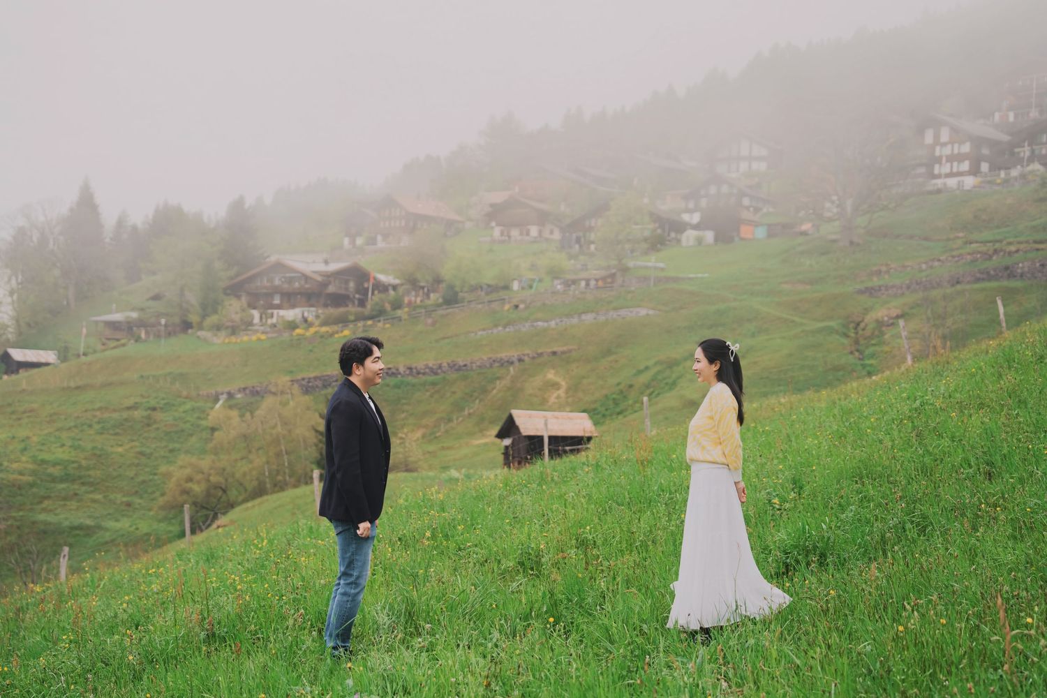 Couple standing apart on a foggy green hillside with traditional Swiss village in the background.