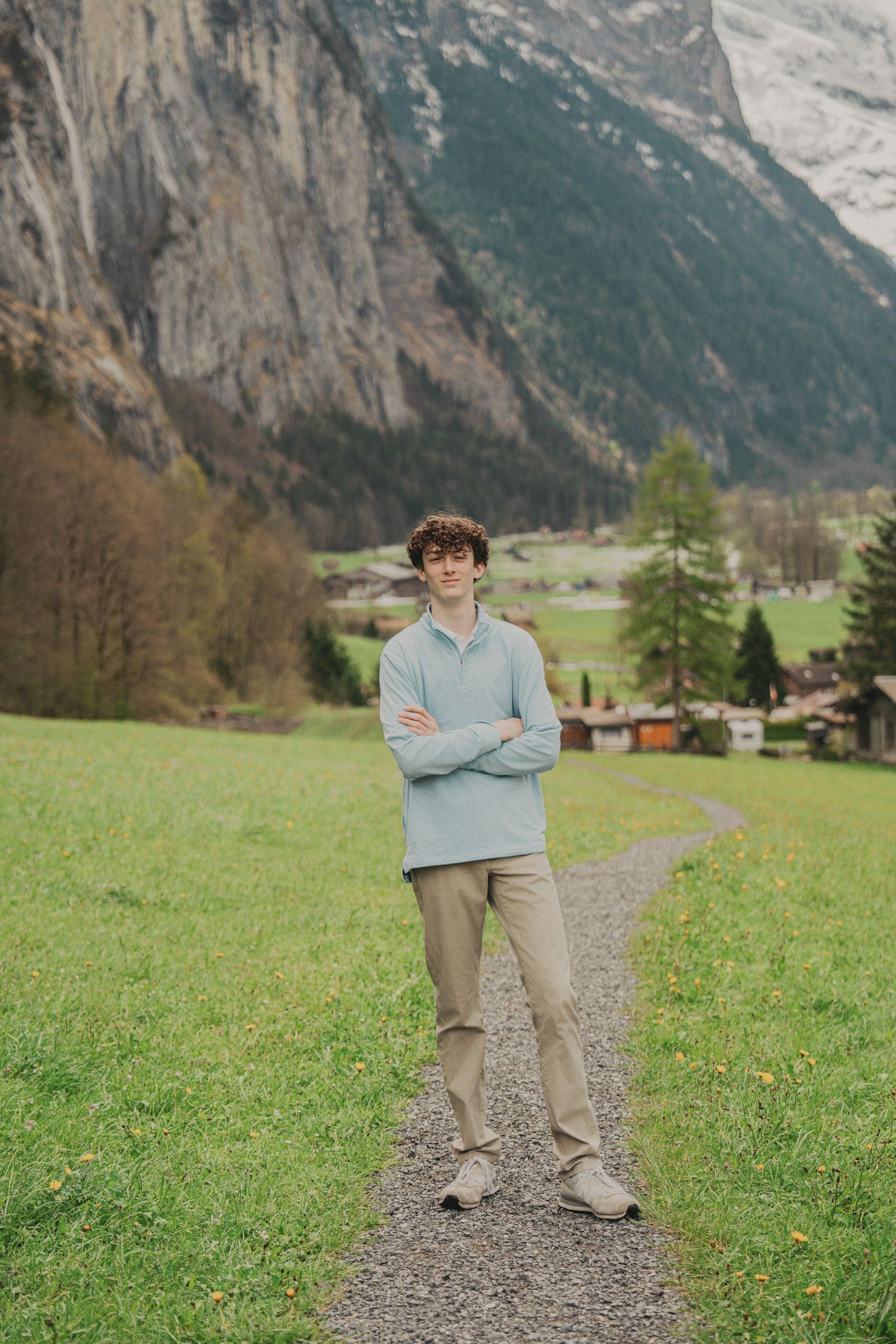 Curly-haired teen in blue pullover standing arms crossed on gravel path in Swiss mountain valley.