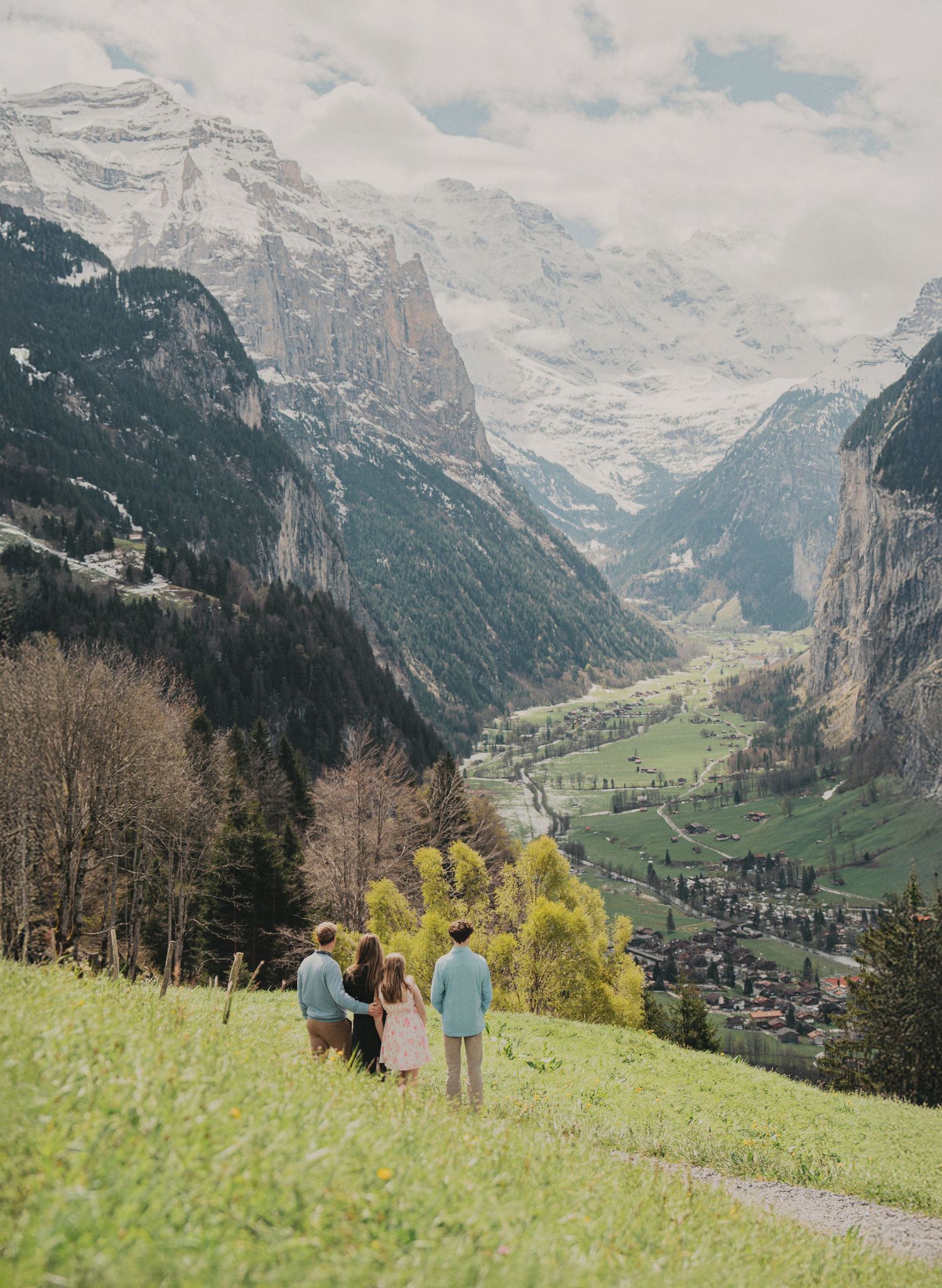 A family of four gazes at a stunning snow-capped Swiss alpine valley from a scenic grassy hillside.