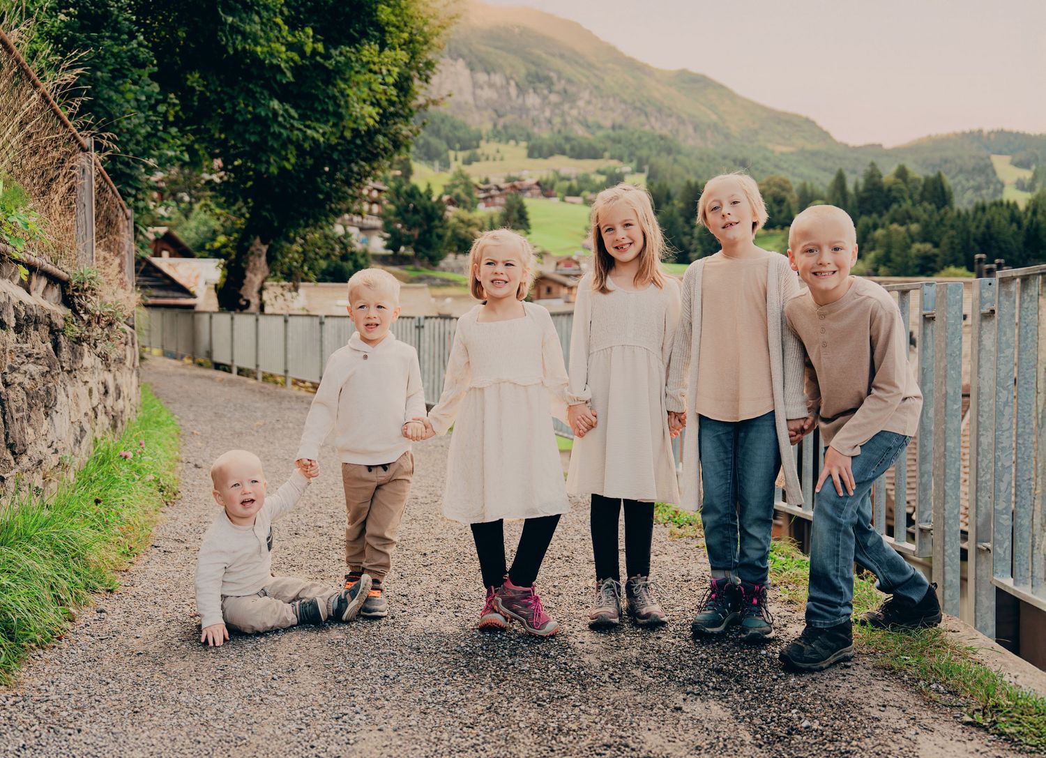 Six young blonde children posing playfully on a gravel path with Swiss alpine mountain scenery behind.