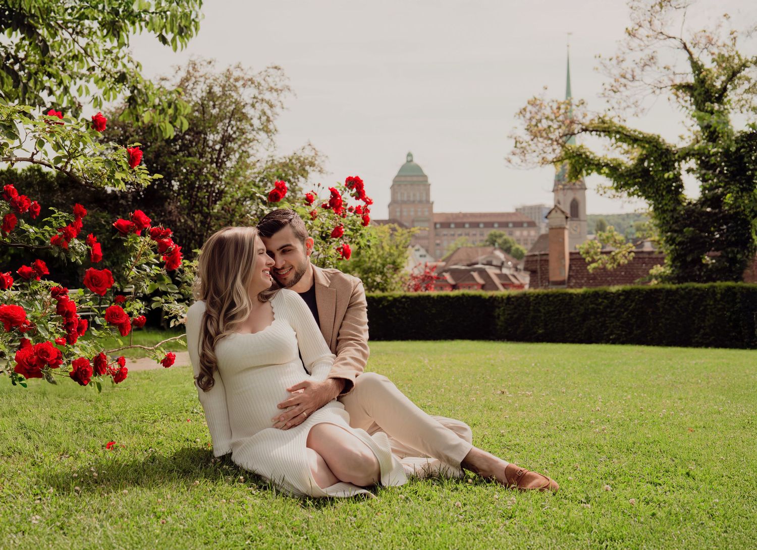 Couple sitting on grass surrounded by red roses, woman in white maternity dress, city skyline behind.