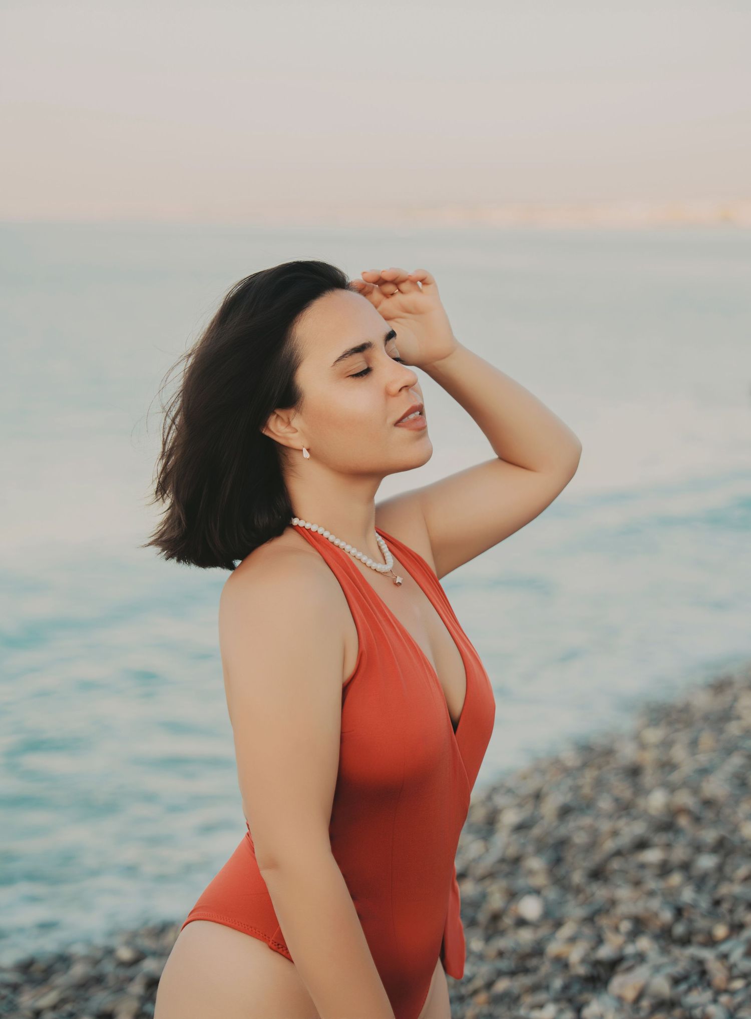 Young brunette woman in red swimsuit shielding eyes from sun on pebble beach at dusk.