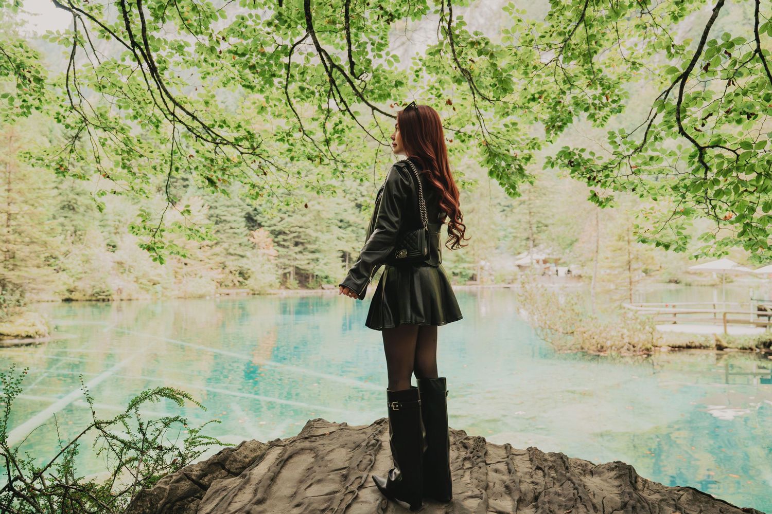 Woman in black leather outfit standing on rocks overlooking brilliant turquoise alpine lake in forest.