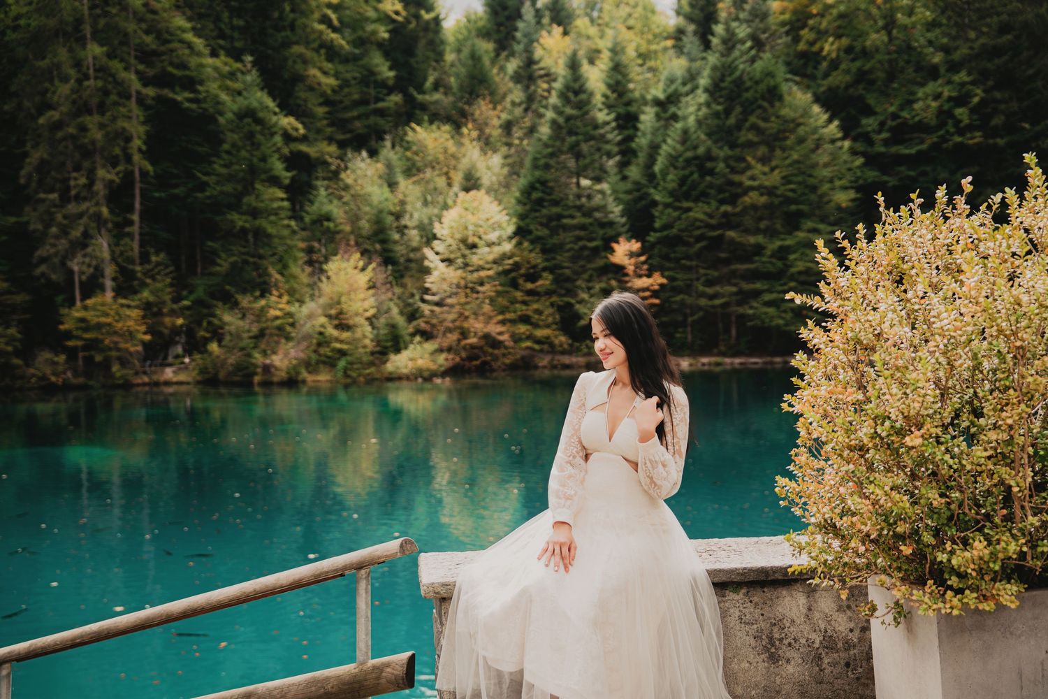 Woman in white lace dress sits pensively on railing by glowing turquoise forest lake.
