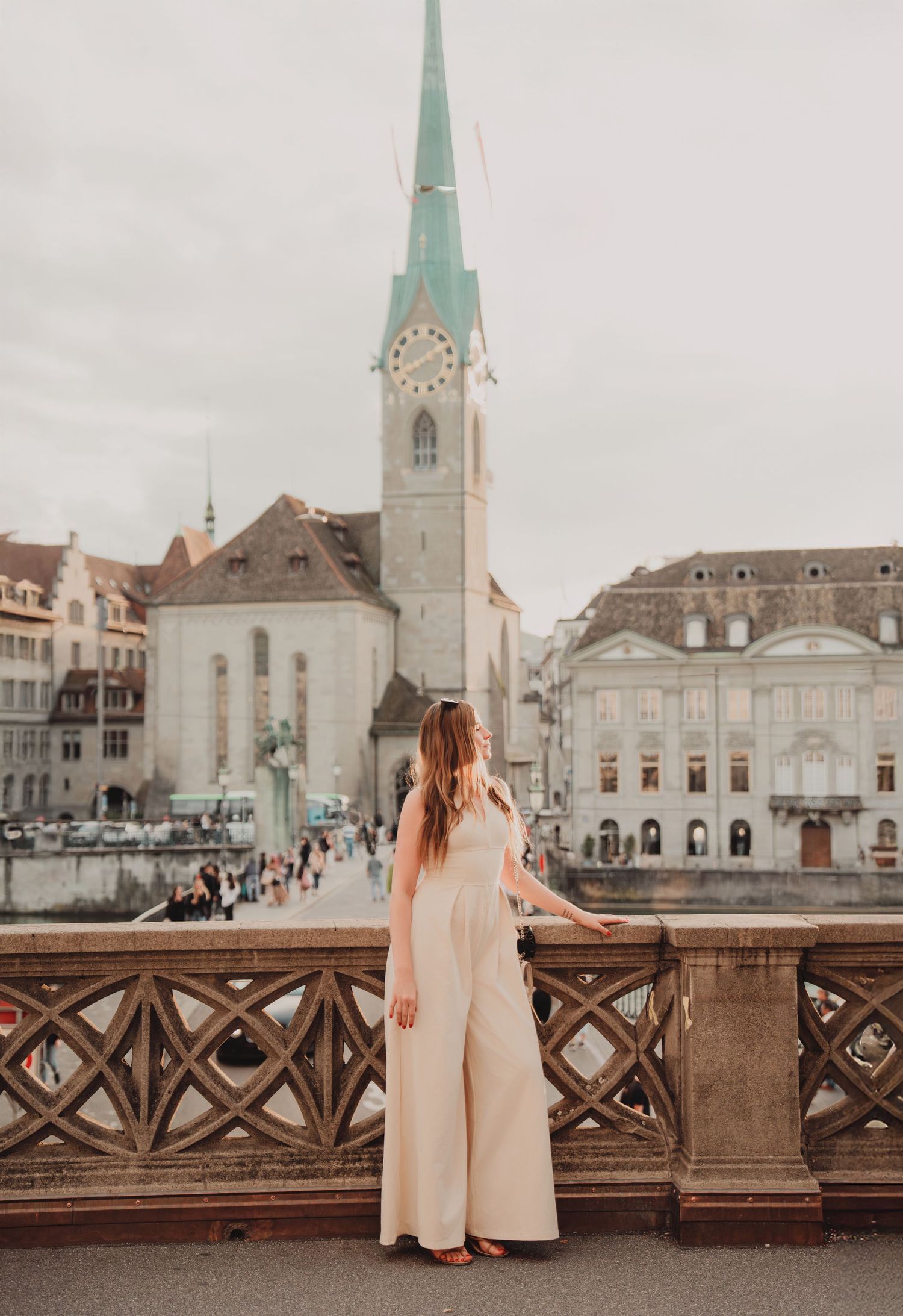 Woman in white outfit standing on historic bridge overlooking Fraumunster church in Zurich oldtown.