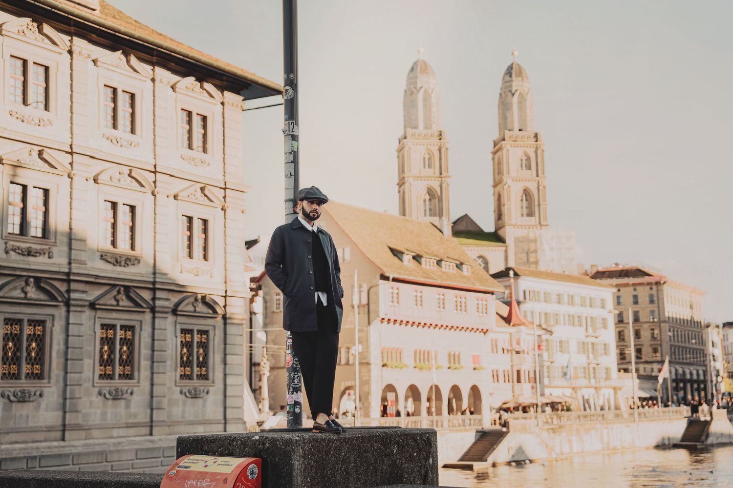 Man in flat cap and dark suit standing on stone platform with Zurich Grossmunster church towers behind.