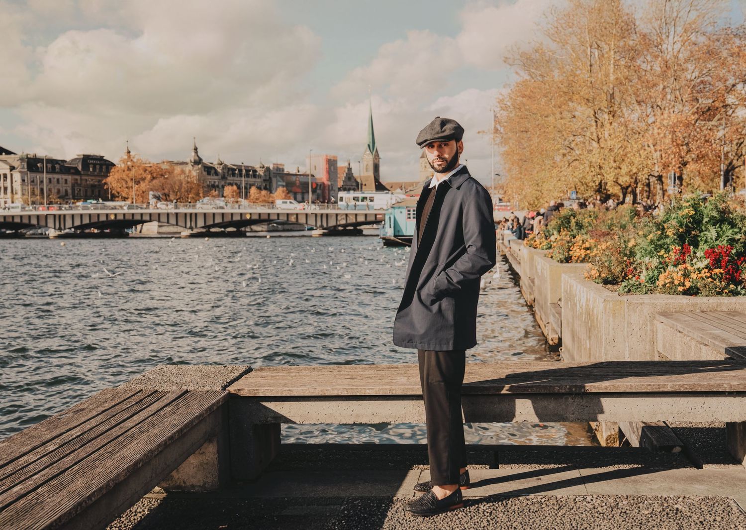 Bearded man in gray flat cap and suit stands on waterfront promenade with historic city skyline in autumn.