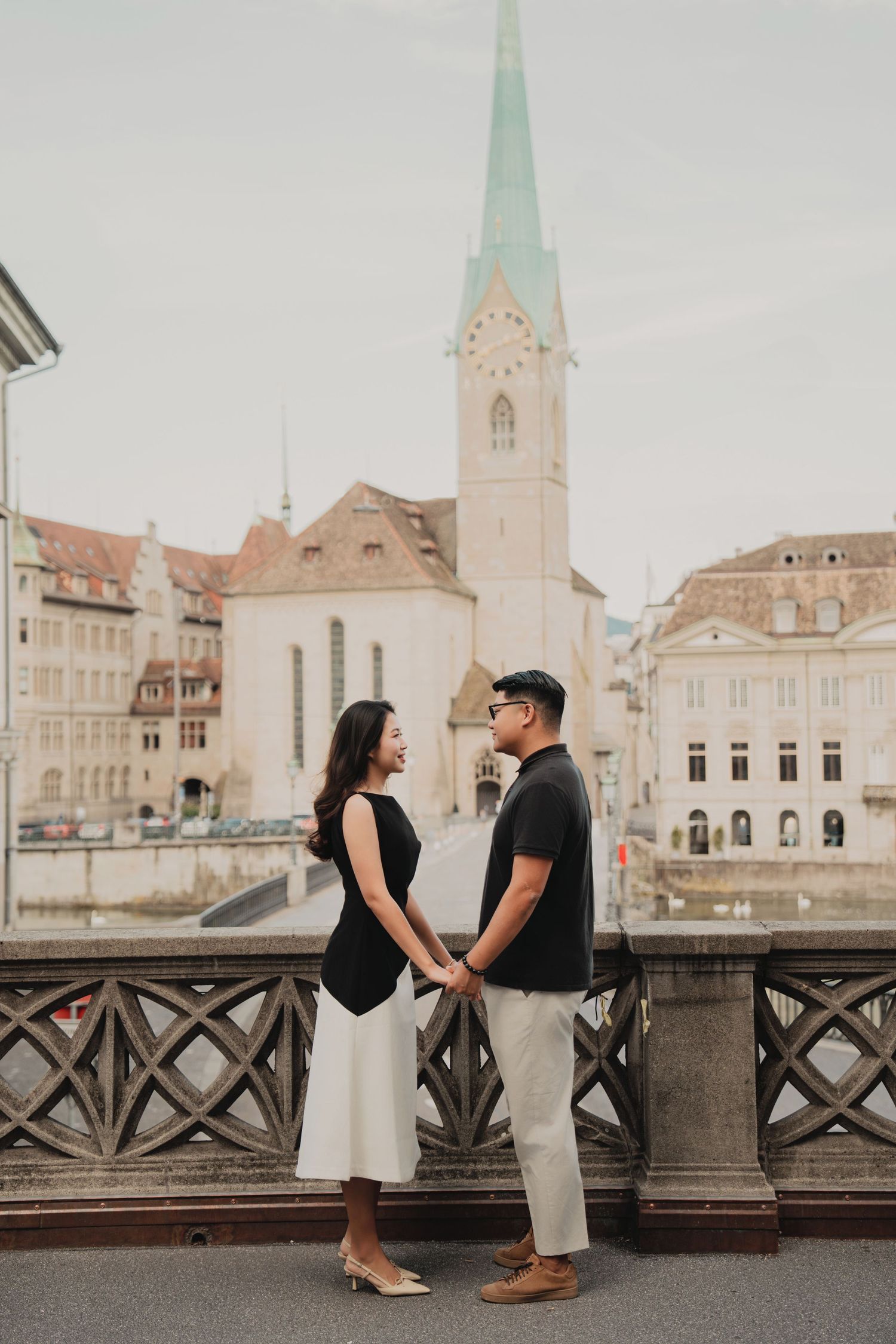 Couple holding hands and facing each other on a bridge in Zurich with church and river backdrop.