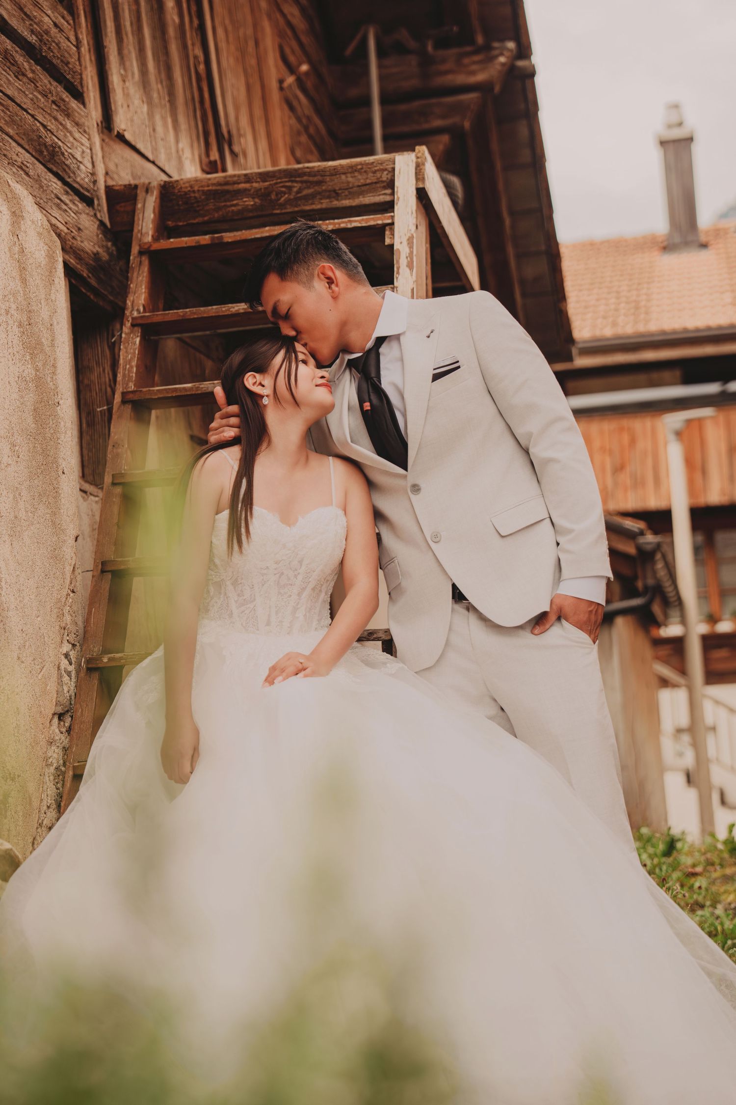 Groom in ivory suit leans in to kiss bride in white ballgown beside a rustic wooden barn.