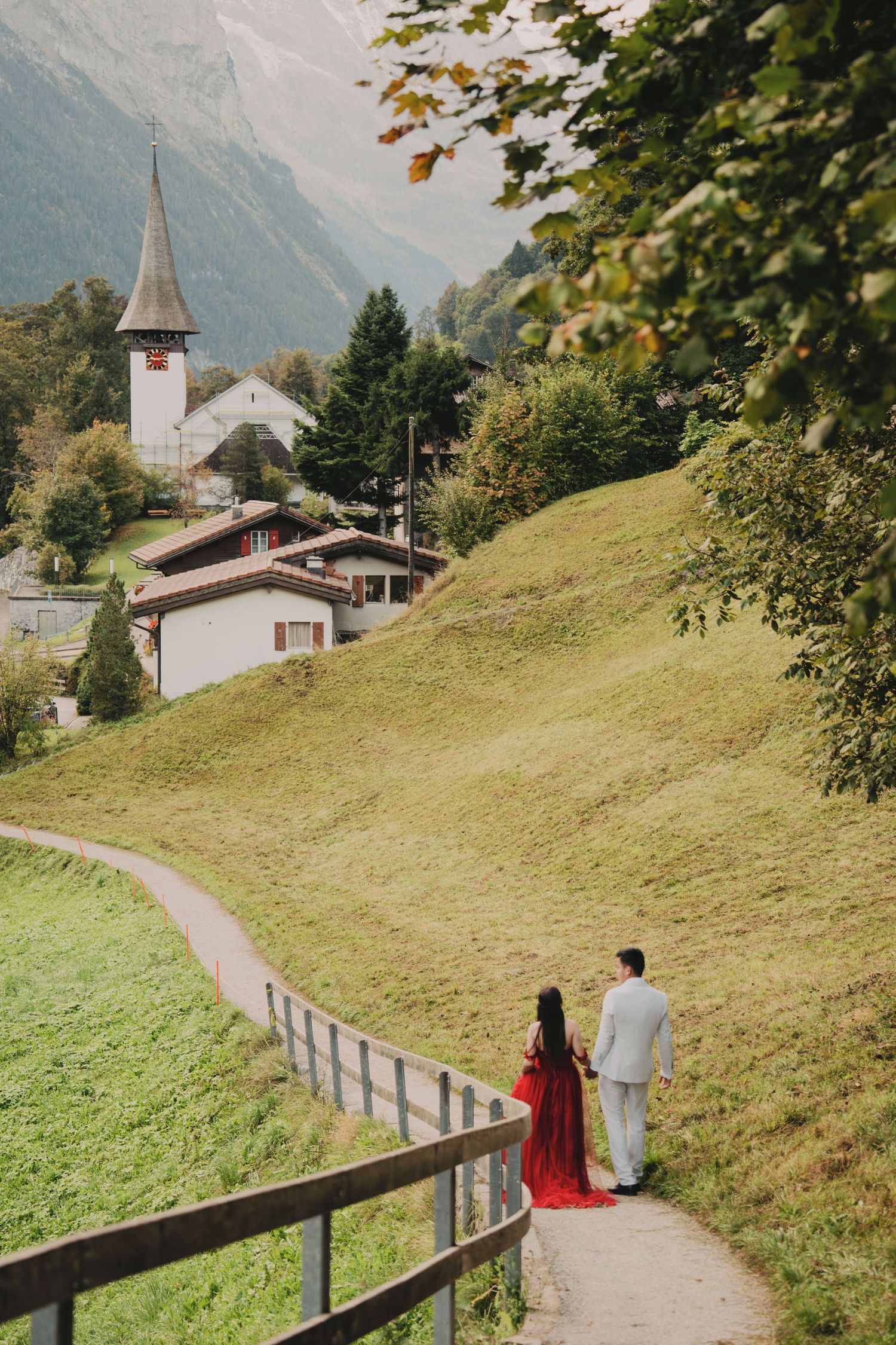 Couple strolling on winding Swiss village path with white church steeple and green hillside.