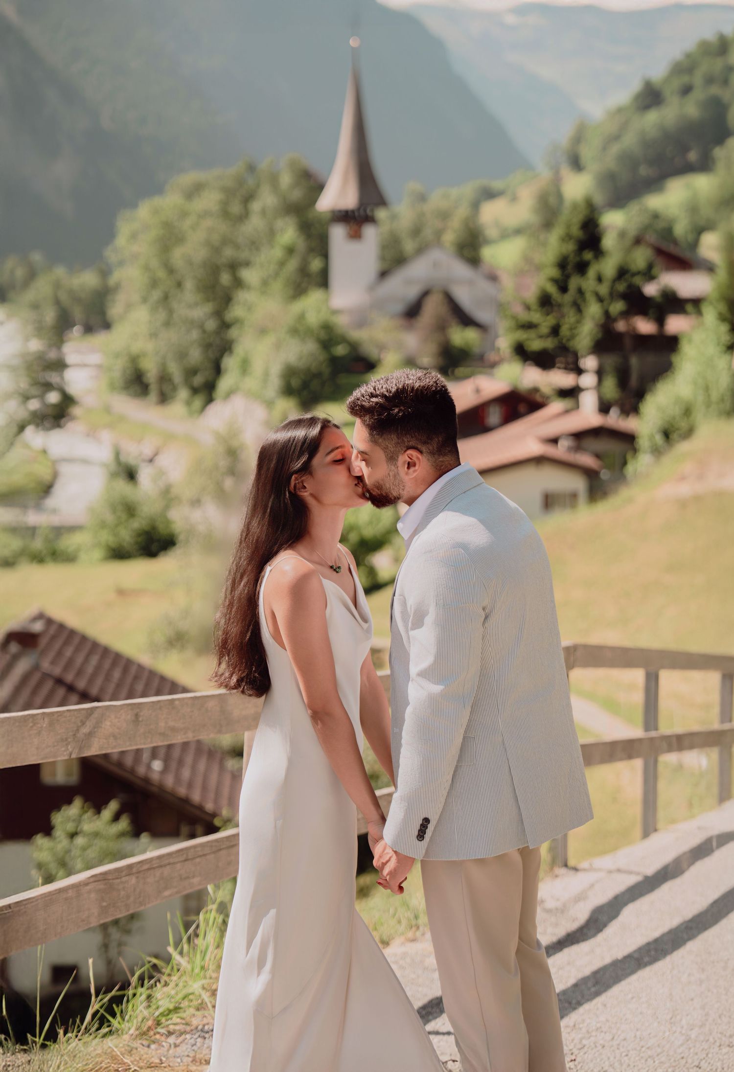 Couple sharing a kiss holding hands with Alpine village church and mountains behind them.