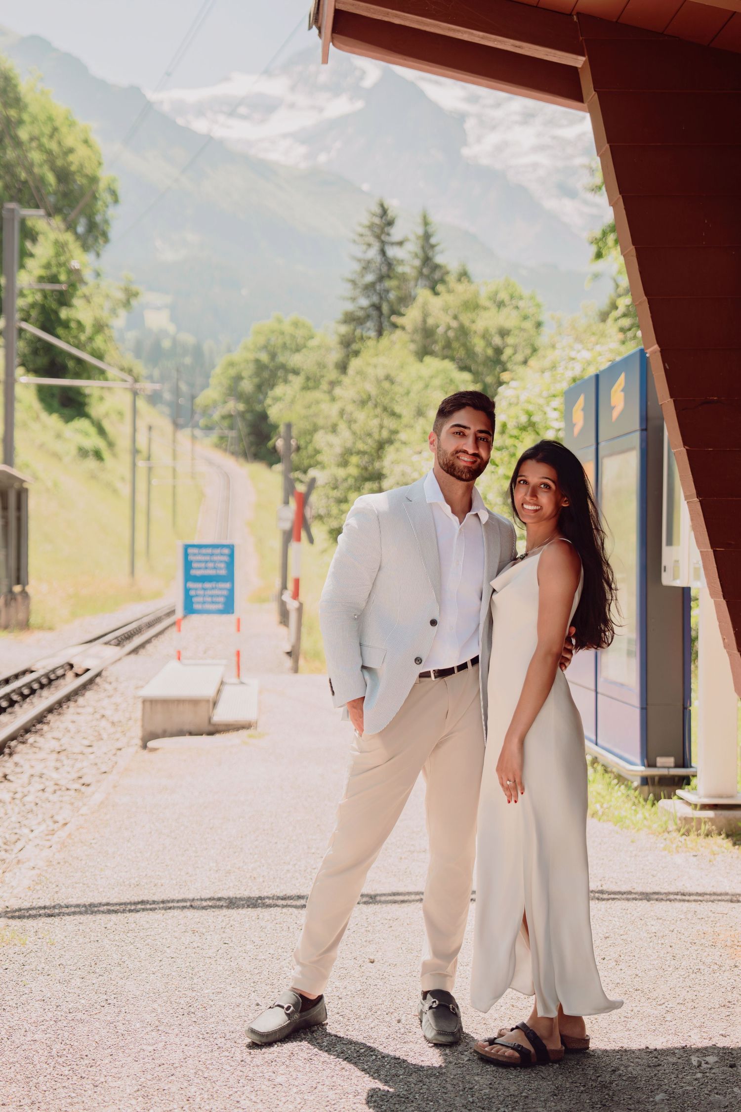 Stylish couple posing together at a scenic Swiss mountain train station with snow-capped Alps in the background.