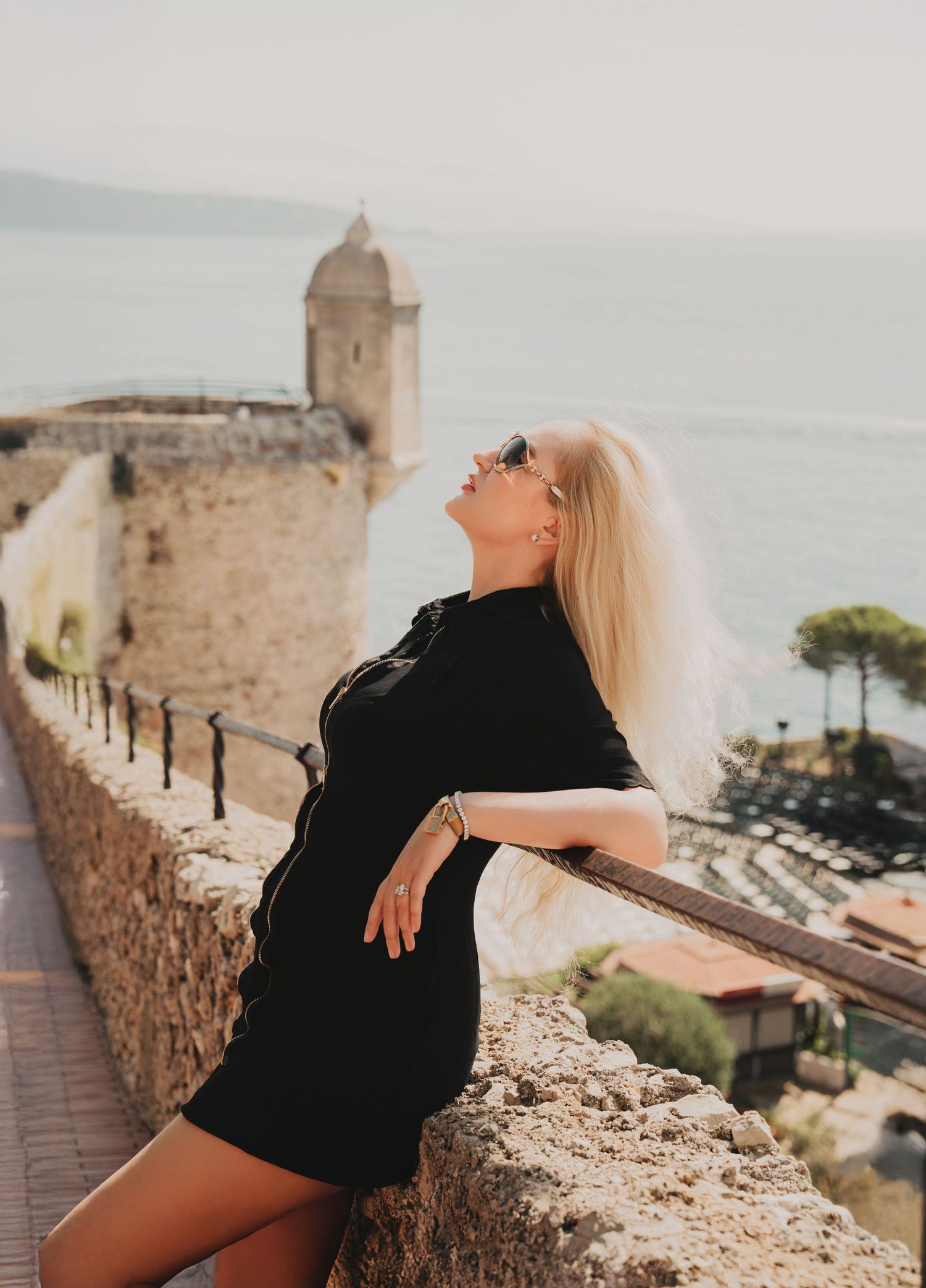 Blonde woman in black dress and sunglasses posing on a scenic coastal fortress wall with ocean views.