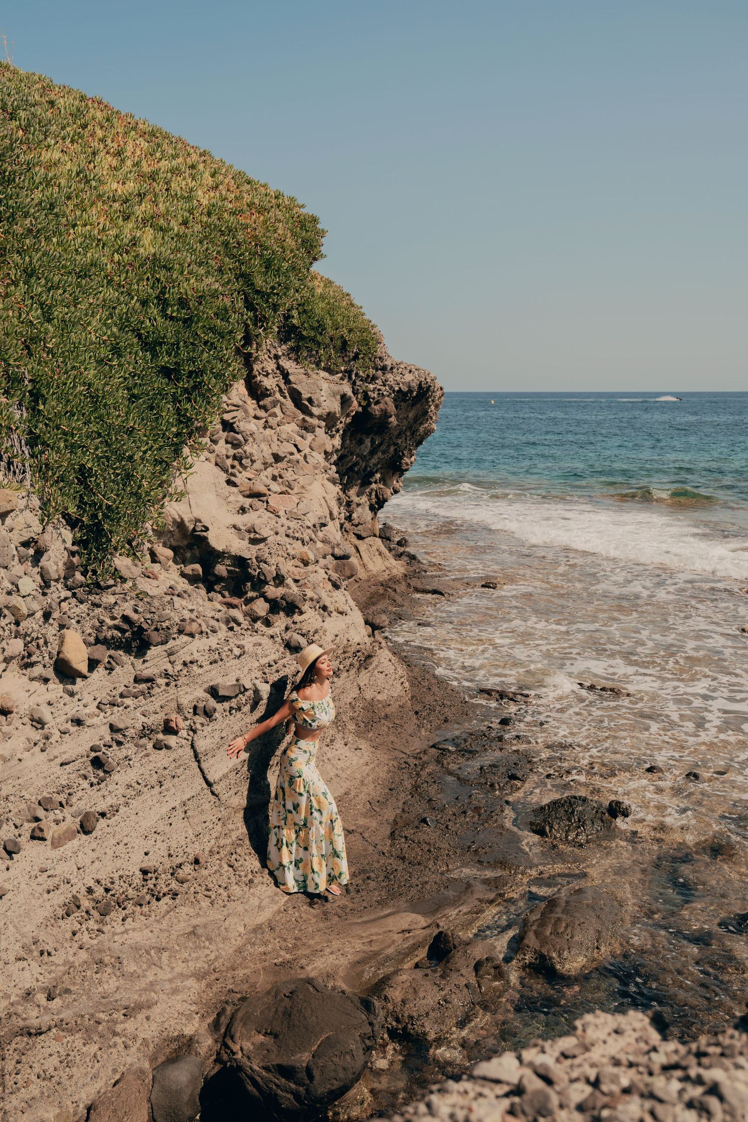 Woman in floral maxi dress standing near rocky coastal cliffs by the blue ocean.