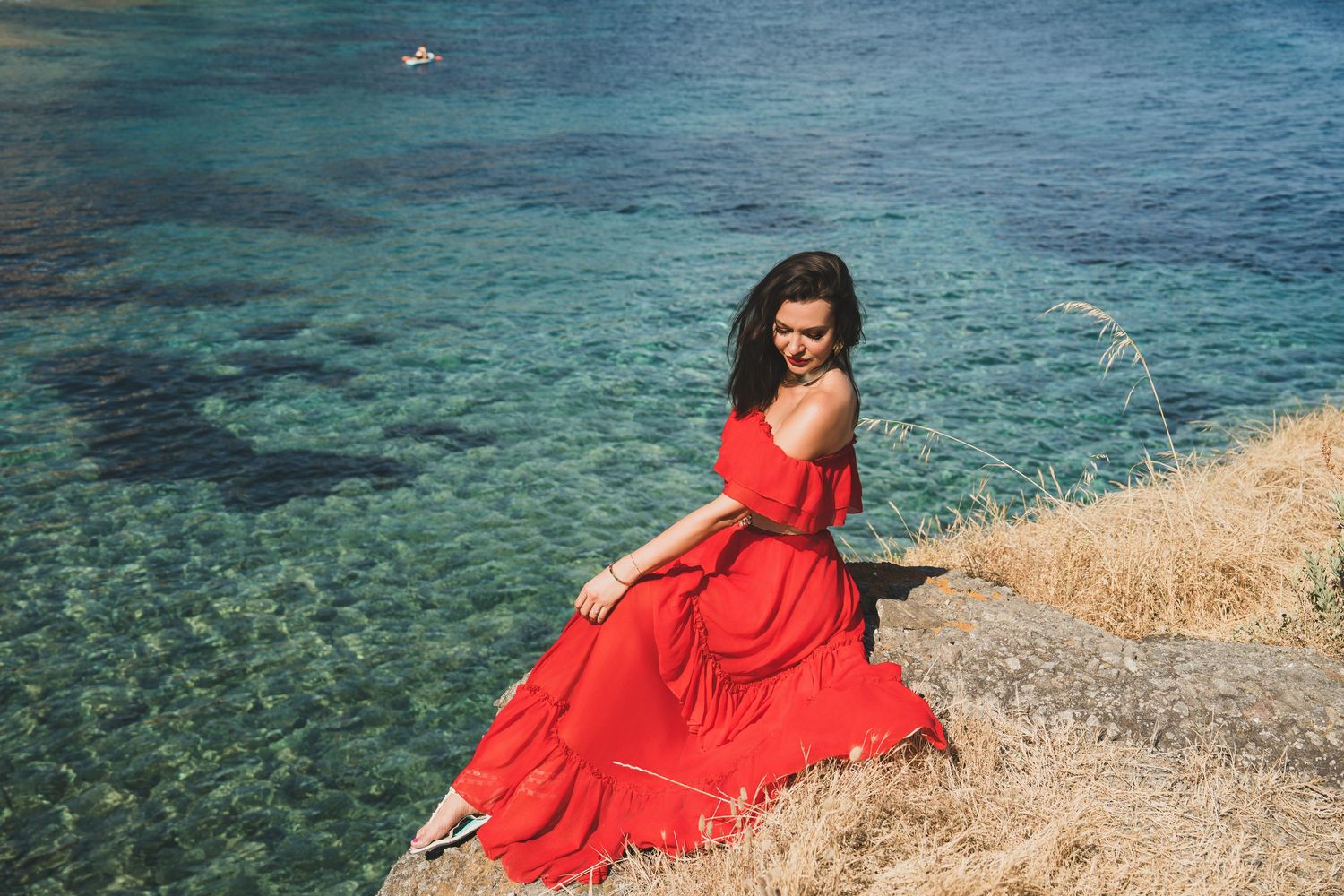 Woman in flowing red dress sitting on rocky cliff edge above clear turquoise coastal waters.