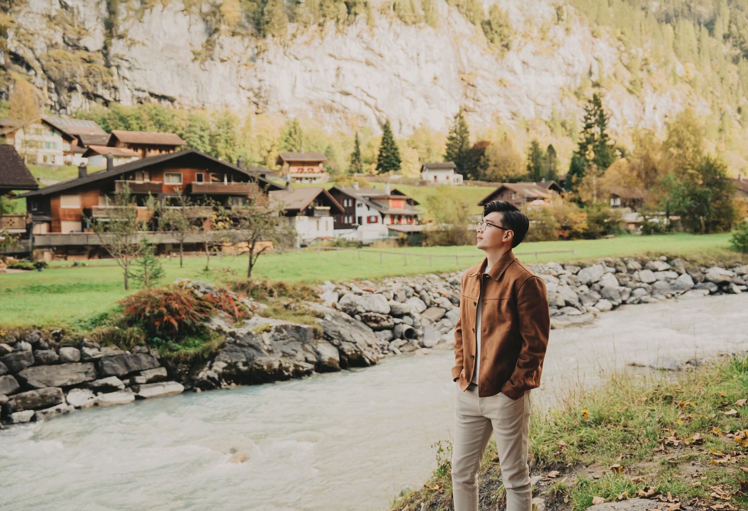 Young man in brown jacket standing beside a rocky mountain river in a Swiss alpine village.