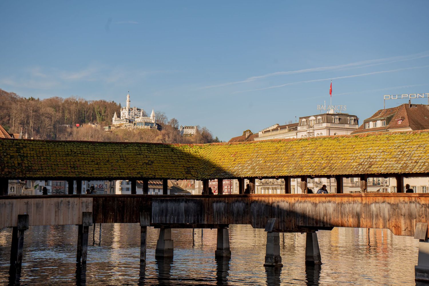 Historic covered wooden bridge over calm water with a hilltop church and blue sky in background.