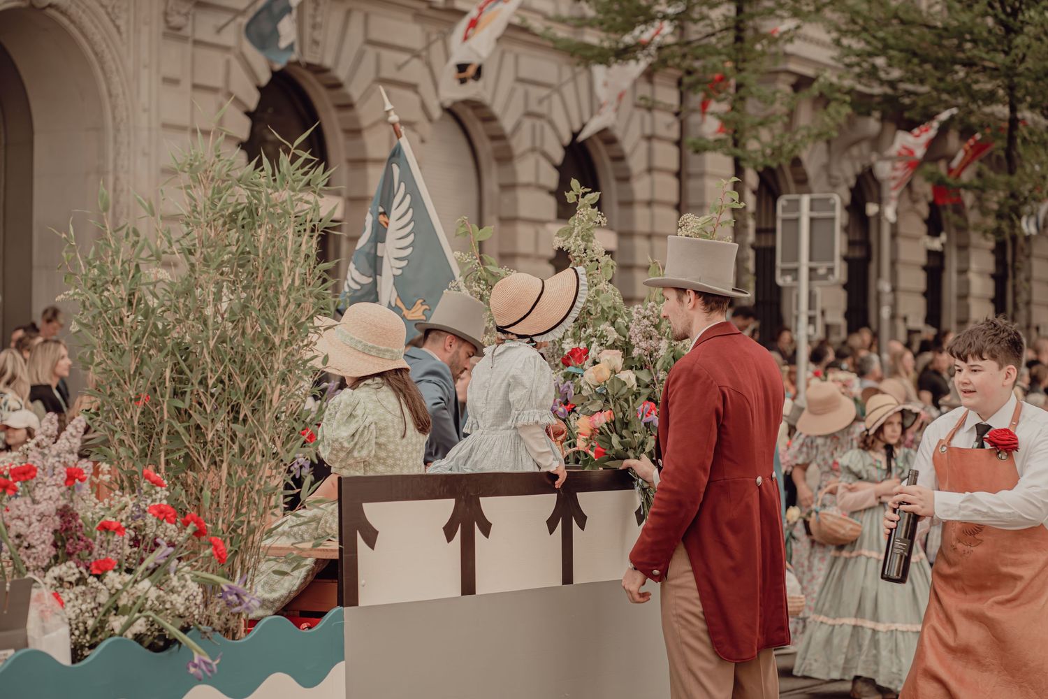 People in Victorian-era costumes standing on a flower-decorated parade float surrounded by colorful blooms and bunting.