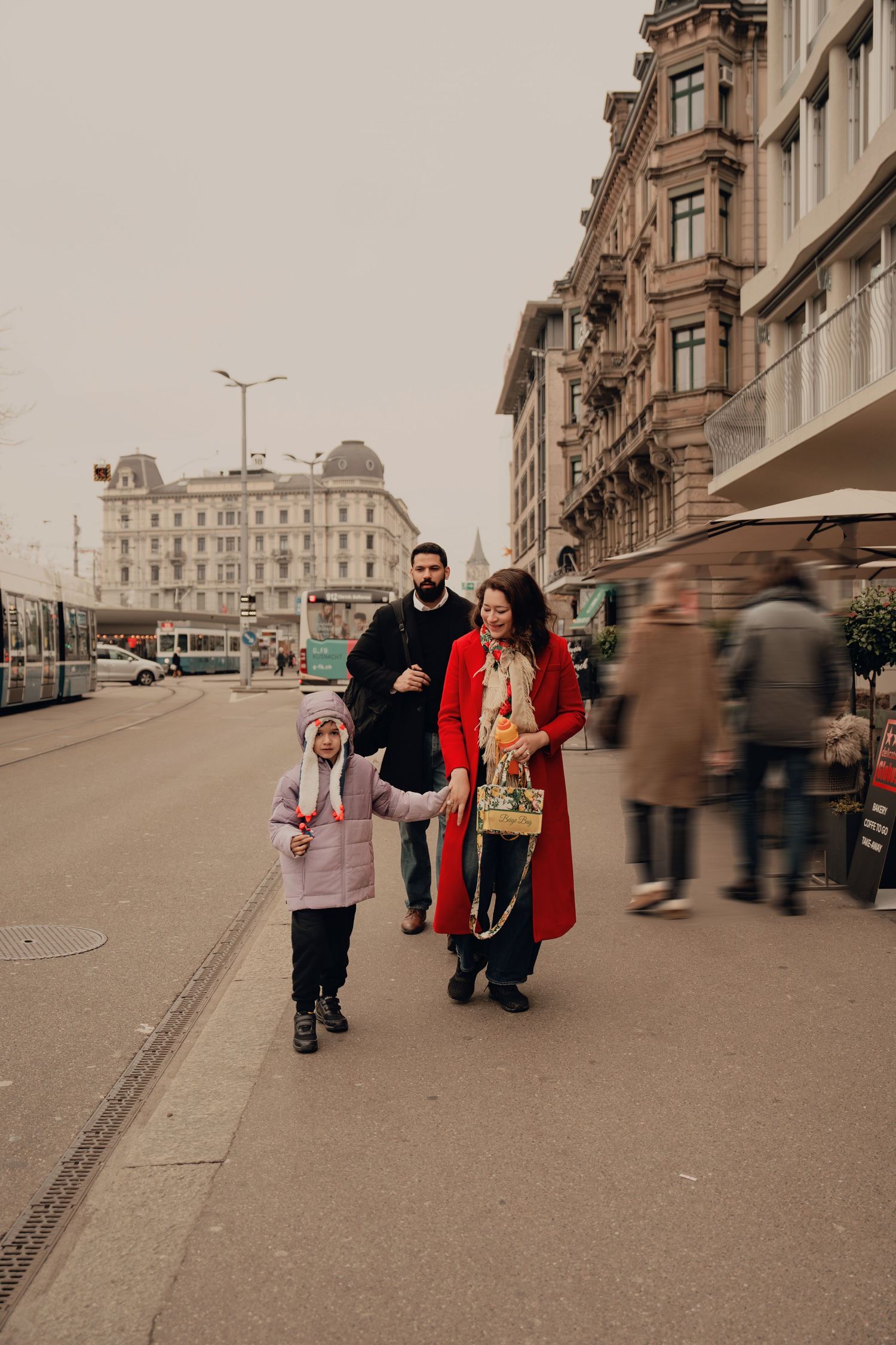 Family of three walking on a city street, woman in red coat holding child's hand, man in black behind.